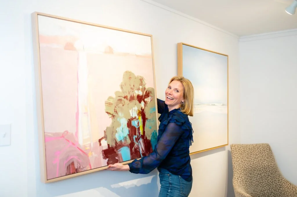 A woman with blonde hair wearing a dark blue blouse and jeans is hanging a painting on a white gallery wall. The painting features a blurred abstract scene with pastel pink, cream, and blue colors. In the background, there is another framed artwork of a seascape and a patterned beige chair.