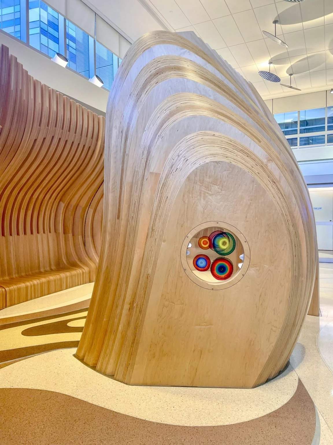 Modern wooden structure with curved layers and colorful circular art installation in an indoor gallery space.