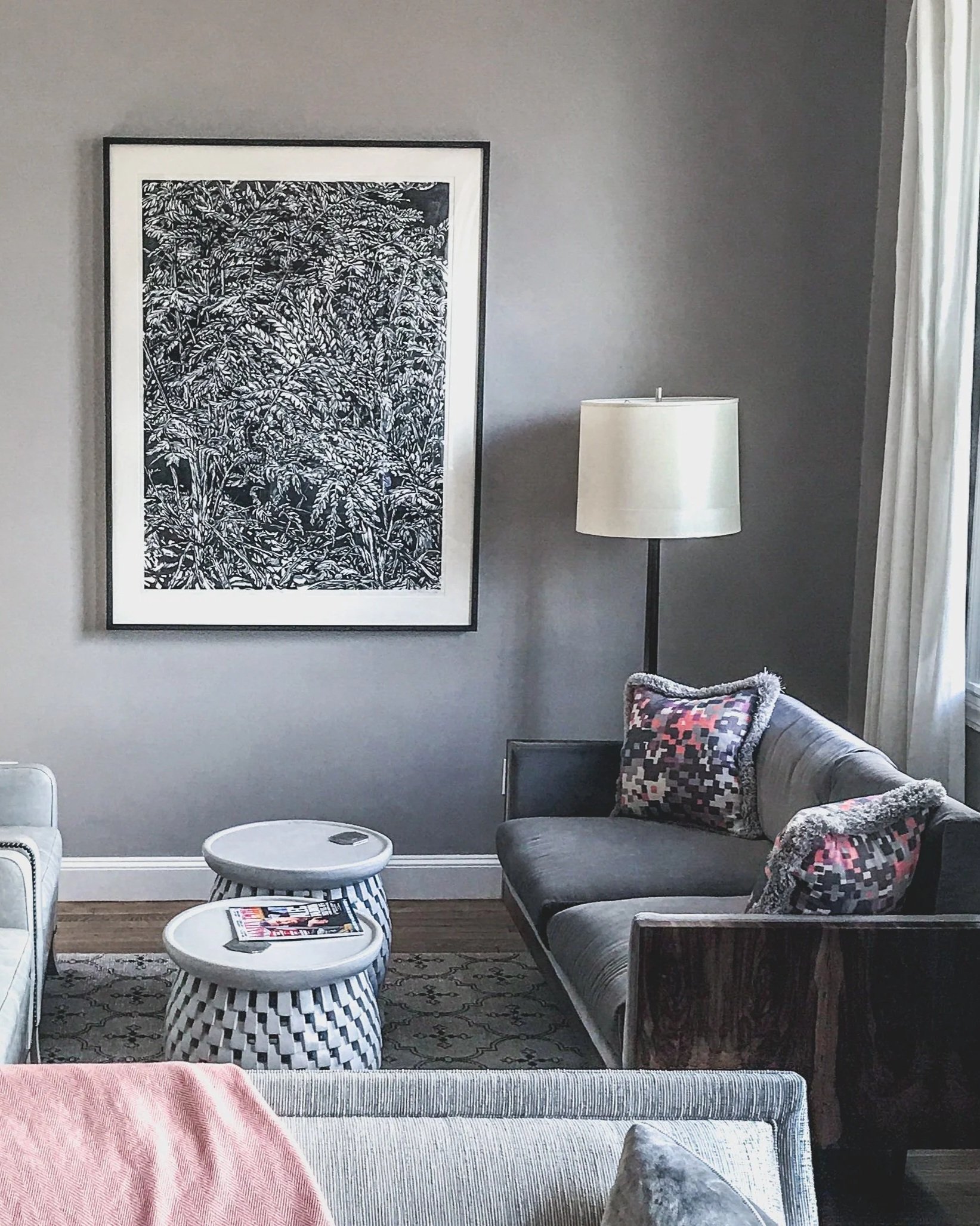 A cozy living room with gray walls, featuring a black-and-white framed artwork, a table with a magazine, a tall lamp, a gray sofa with patterned pillows, and a draped curtain next to a window.