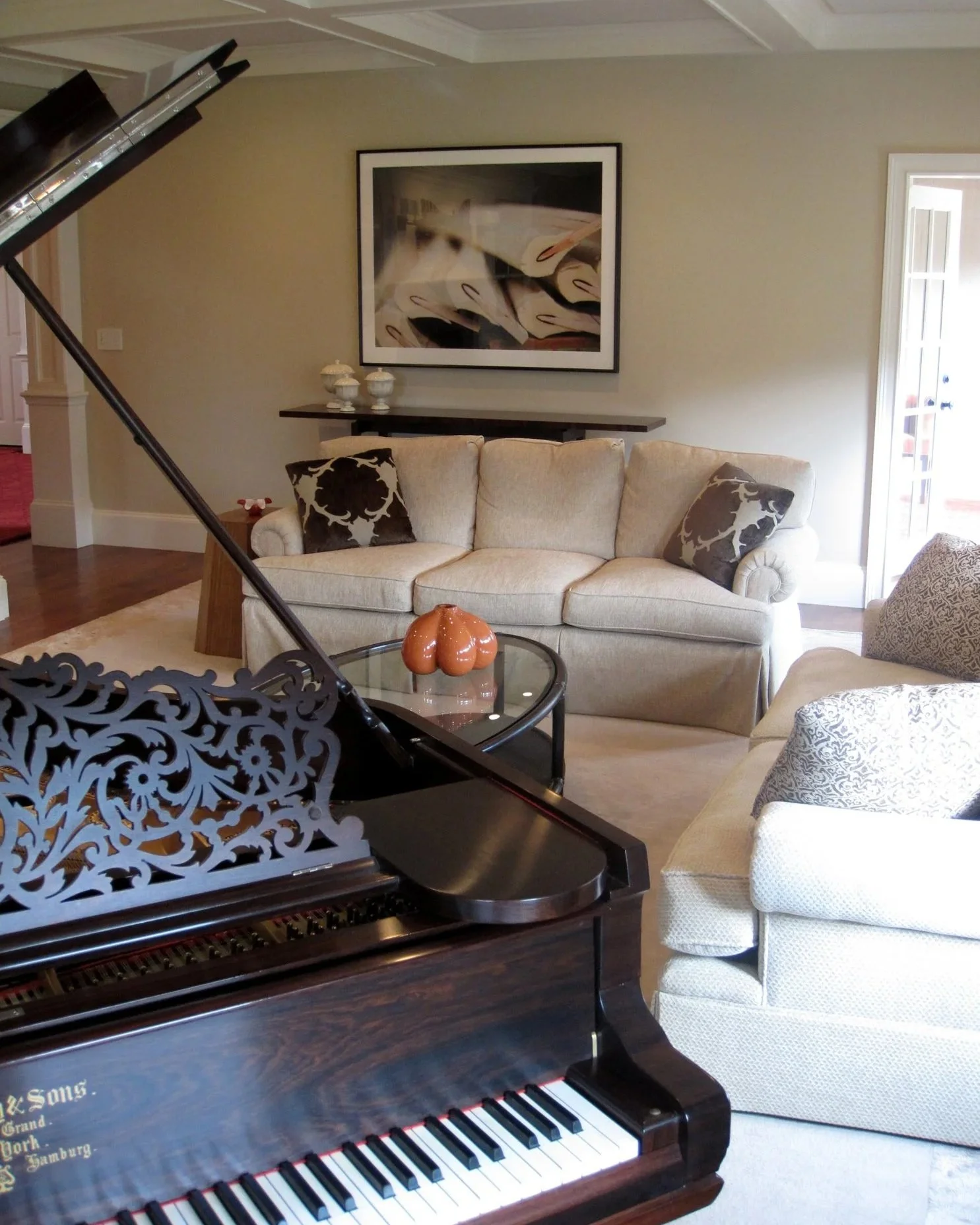 Living room with a grand piano, a beige sofa with decorative pillows, a framed abstract art piece on the wall, a small table with a pumpkin-shaped decoration, and a door leading outside.