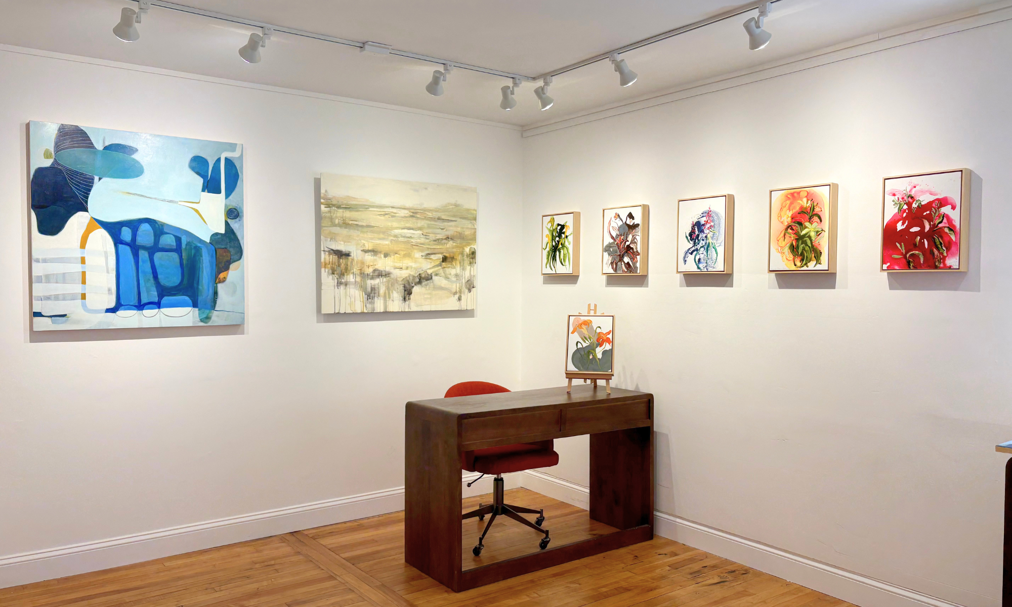 An art gallery wall displaying various abstract and floral paintings, with a small easel holding a framed artwork on a wooden table in front of the wall.