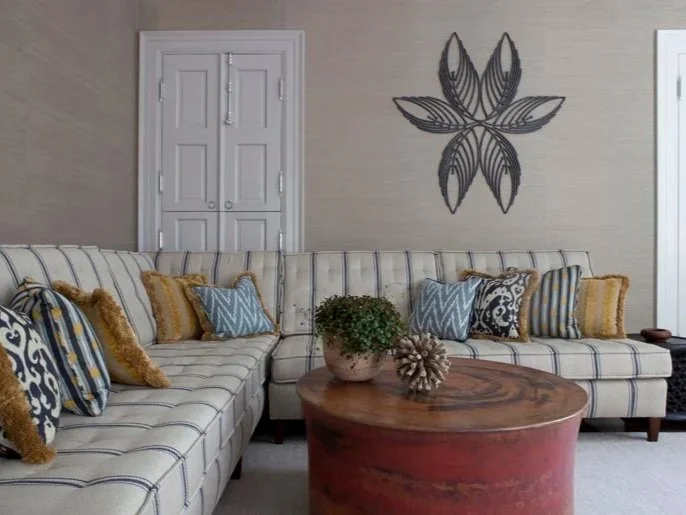Living room with a large striped sectional sofa, decorative cushions, a round wooden coffee table with a potted plant and decorative item, beige walls, a wall art piece, and double doors in the background.