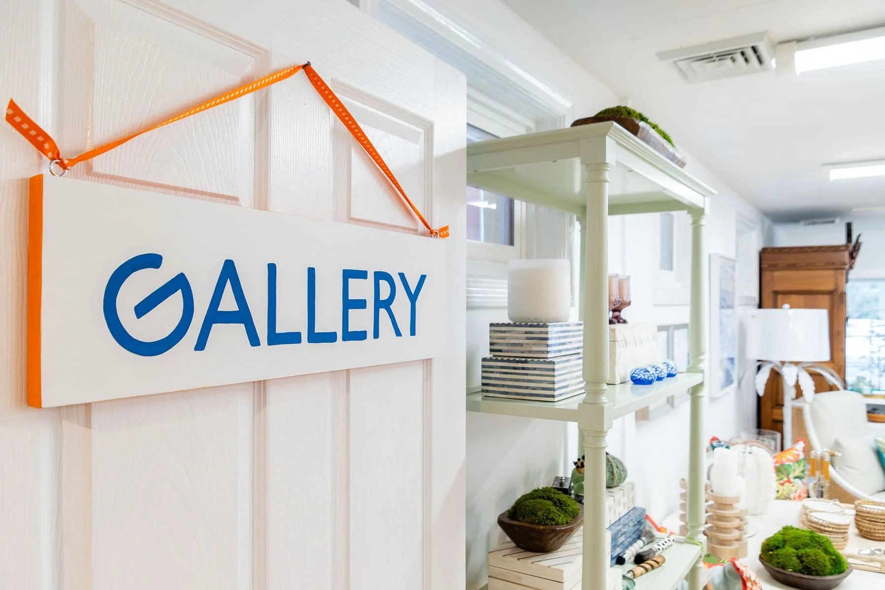 A white door with a sign that says 'GALLERY' in blue lettering, hanging with an orange strap in a shop filled with decorative items such as baskets, pots, and plants.