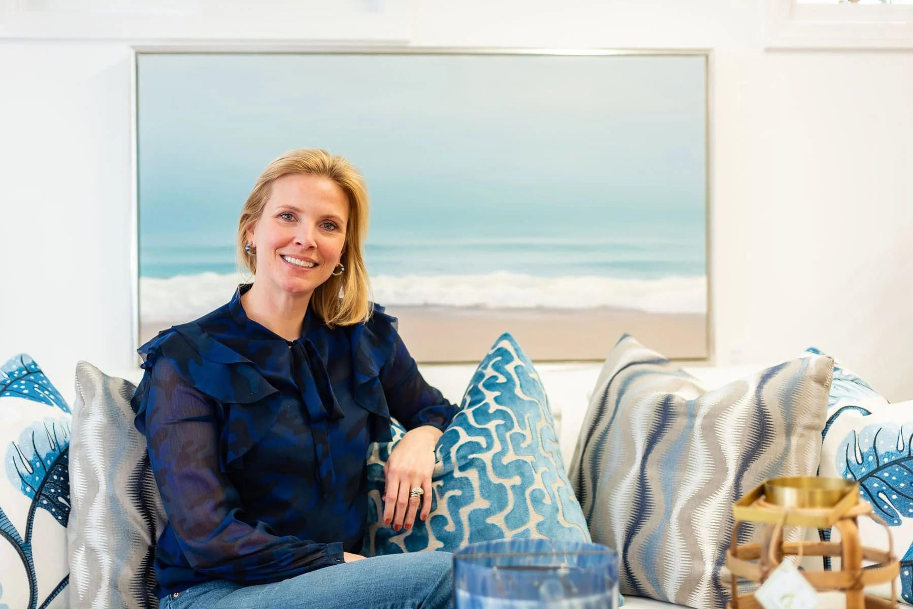 A woman with blonde hair and a blue patterned blouse sitting on a sofa with blue and beige patterned cushions, by a large window showing the beach and ocean.