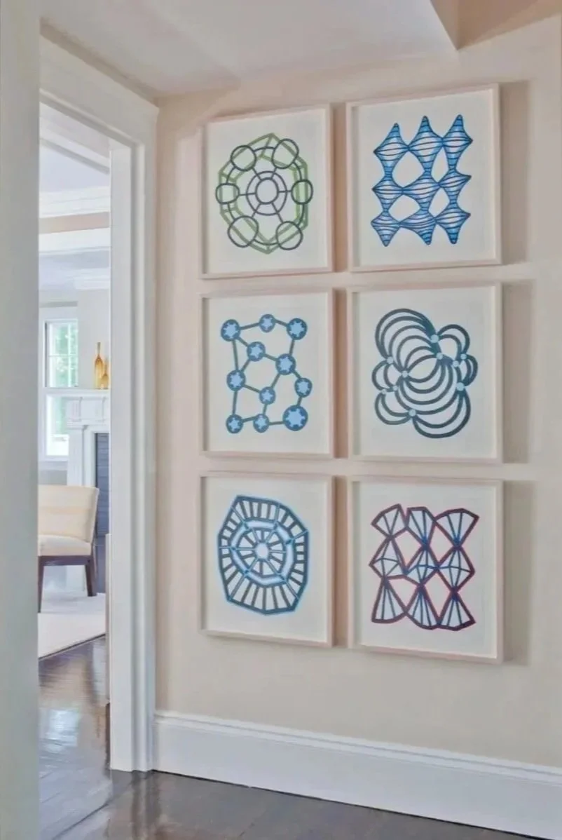 Six framed abstract geometric line art prints in blue, green, and red, arranged in two columns and three rows on a white wall in a home interior.