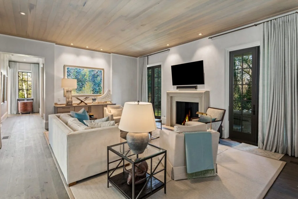 Living room with beige sofas, armchairs, a fireplace, wall-mounted TV, large windows with curtains, a wooden ceiling, and a view of trees outside.