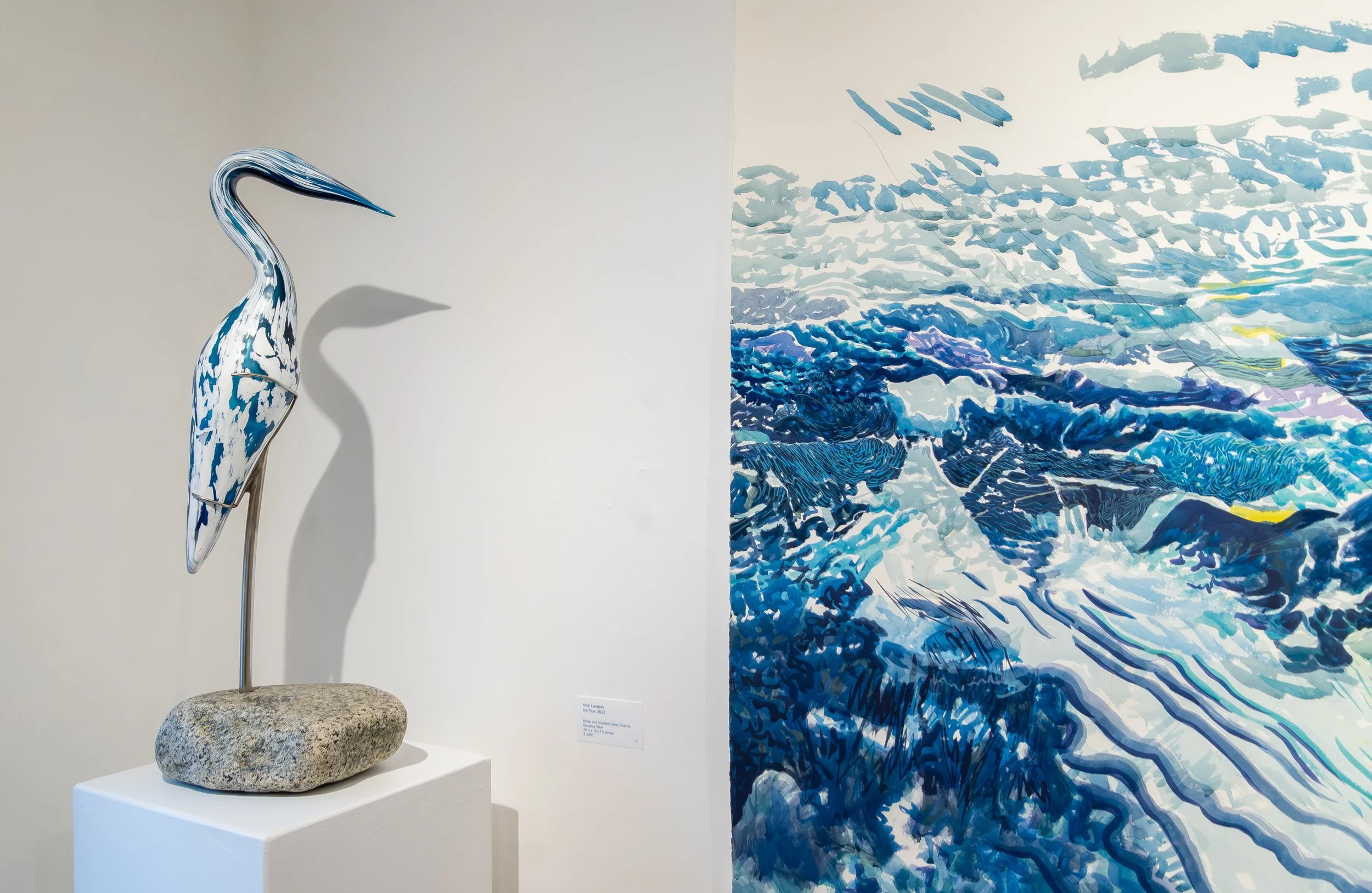 A sculpture of a heron or egret, painted with a blue and white pattern, stands on a stone base. Next to it, an abstract painting in various shades of blue depicts water and sky.
