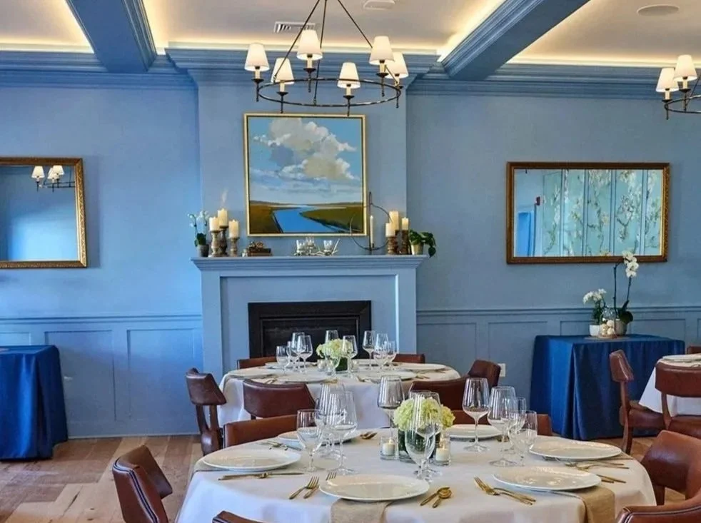 Elegant dining room with round tables covered in white tablecloths, set with plates, glasses, and gold utensils, decorated with white flowers. The room features blue walls, chandeliers, mirrors, a fireplace, and wooden flooring.