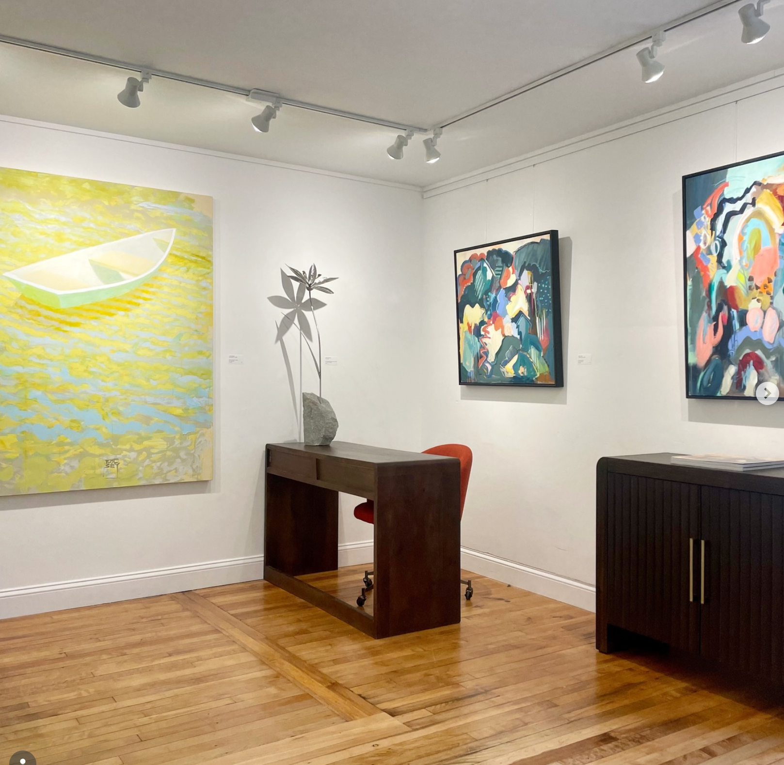 Interior of an art gallery displaying colorful paintings on white walls, wooden flooring, and a dark wooden table with an orange chair, a sculpture of a plant on a stone base.