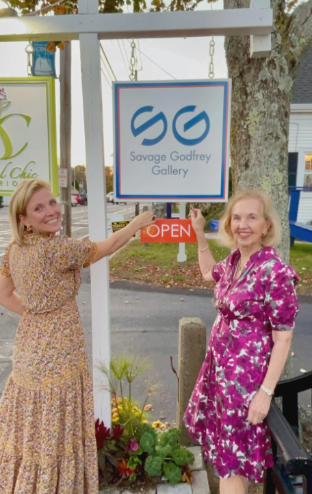 Two women standing outside in front of a gallery sign that reads "Savage Godfrey Gallery" with an "OPEN" sign, both smiling and holding up the sign.