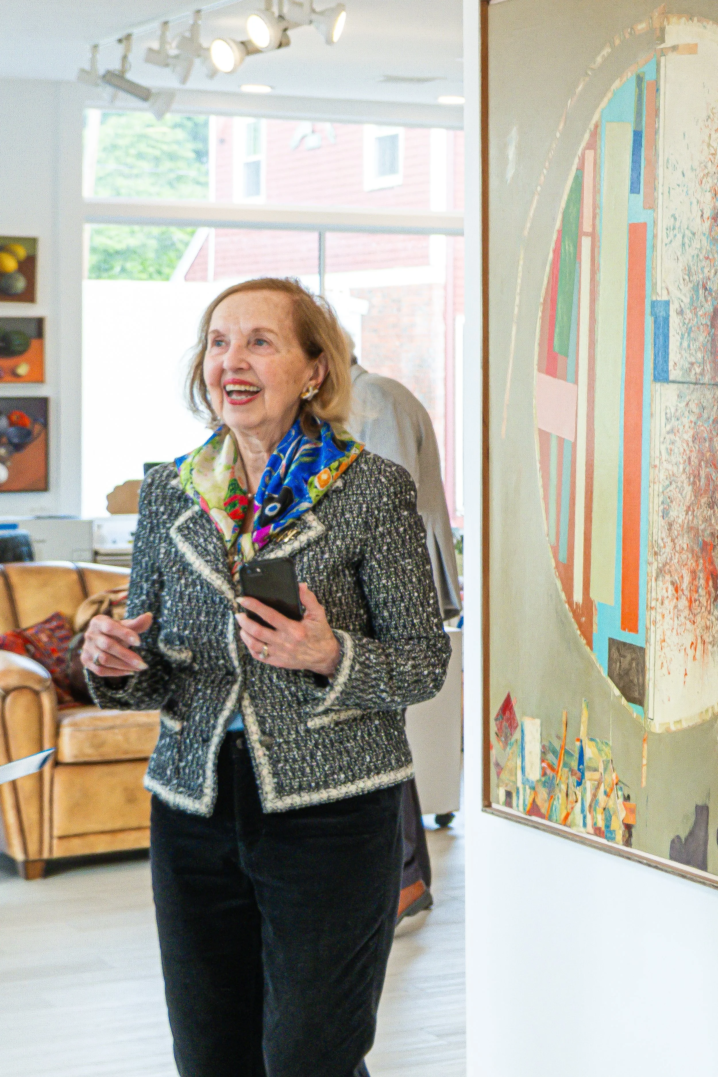 An elderly woman with light brown hair smiling, dressed in a black and white patterned blazer and a colorful scarf, holding a smartphone in her right hand. She is standing in an art gallery or museum, with artwork on the walls and a wooden chair visible in the background. Large windows let in natural light.