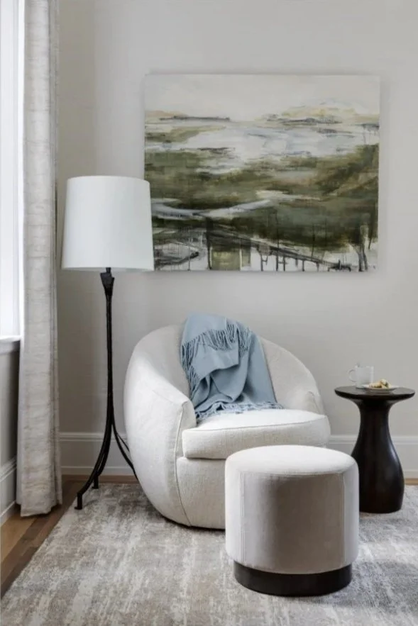 A cozy living room corner with a white upholstered armchair, a matching ottoman, a tall floor lamp, a side table with a cup and snack, and a large abstract landscape painting on the wall.