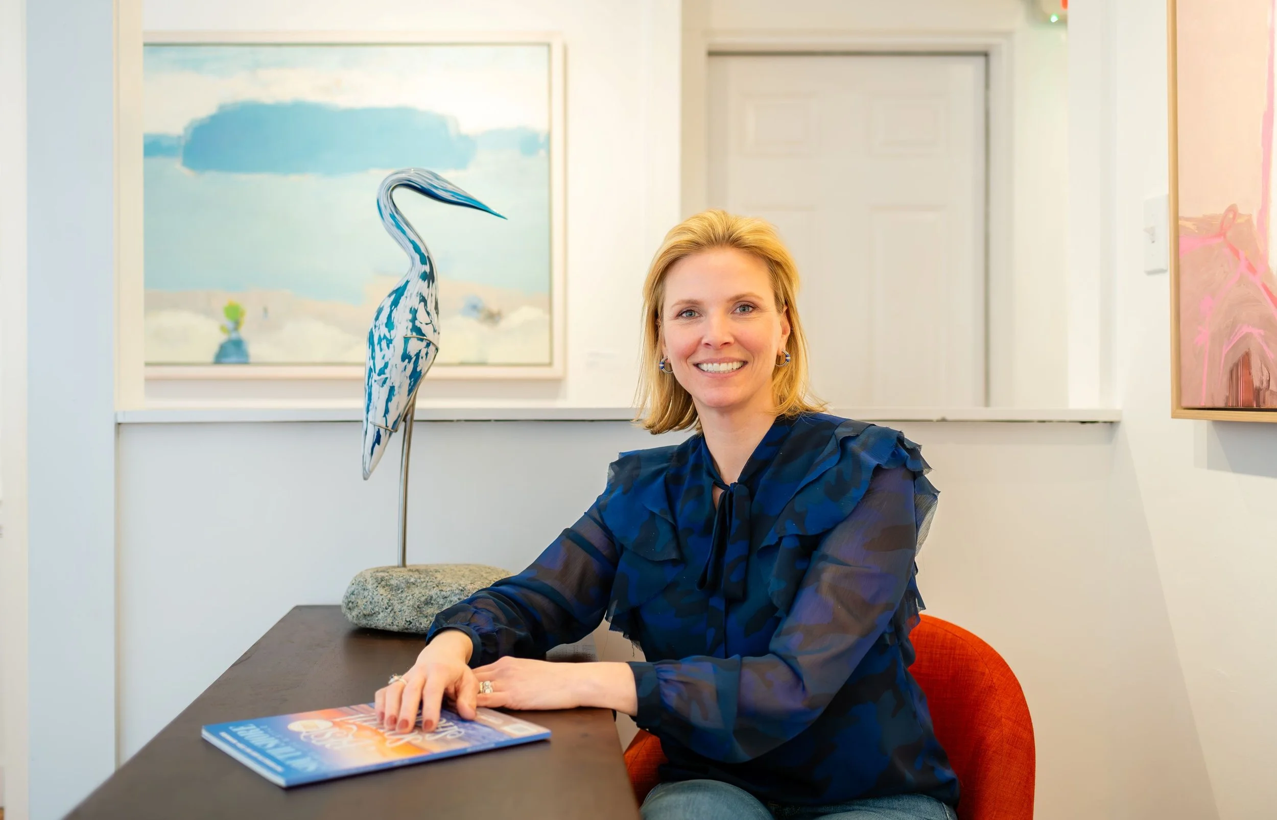 A woman with blonde hair and blue eyes, wearing a dark blue blouse, sitting at a table with a magazine, smiling. Behind her are colorful paintings and a framed artwork of a crane sculpture on a stone base.