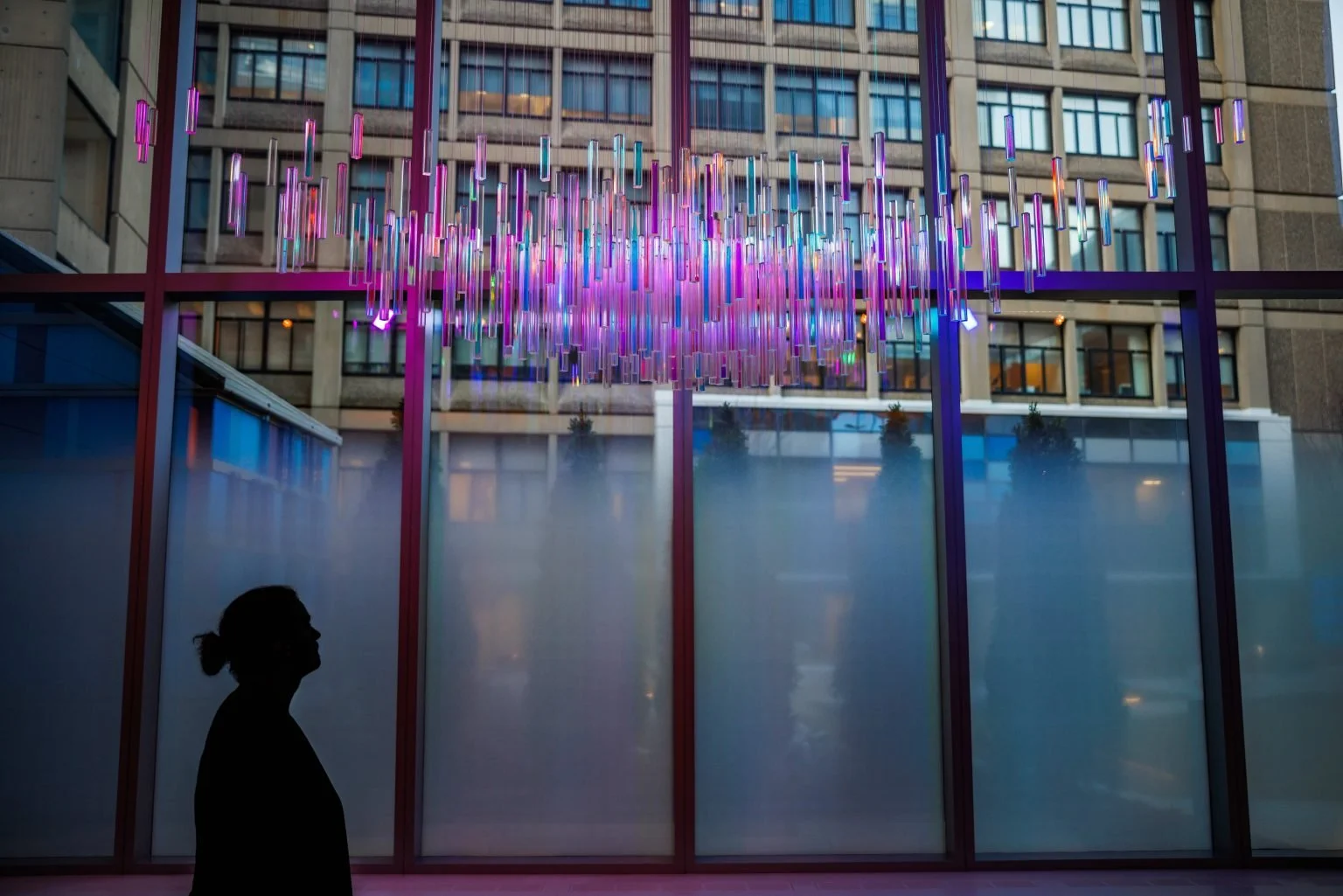 Silhouette of a person with a bun hairstyle looking at a modern, colorful hanging light installation outside a building with glass windows.