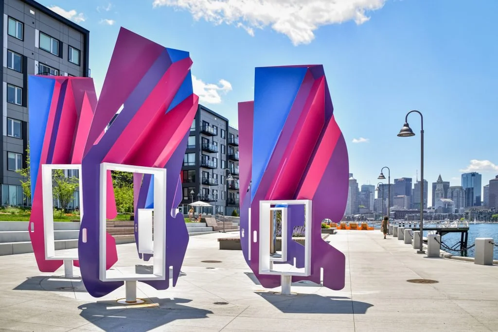 Colorful outdoor art installation with pink and purple abstract shapes and white frames on a city waterfront promenade under a blue sky.