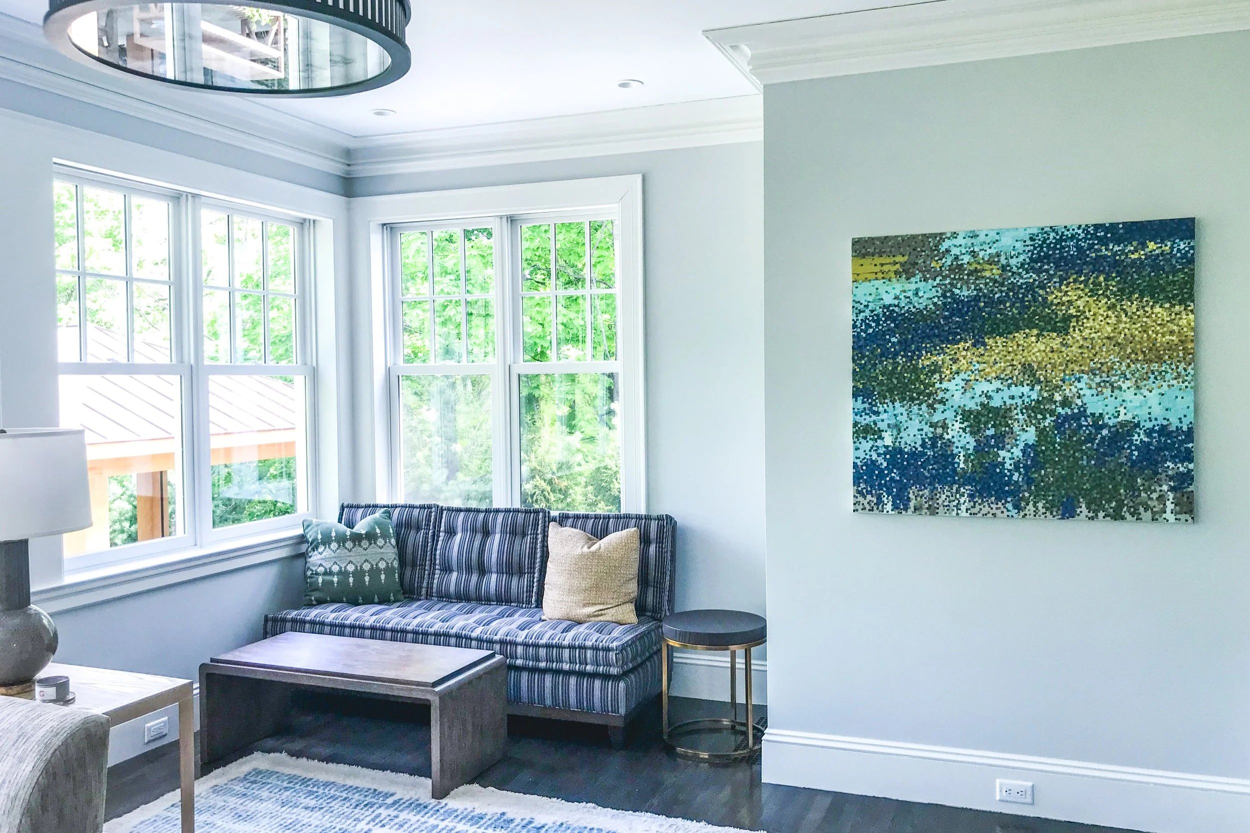 A living room with large bay windows, a blue and white striped sofa with decorative pillows, a wooden coffee table, a small round side table, a white desk lamp, a framed abstract painting on the wall, and a blue and white patterned rug on a dark hardwood floor.