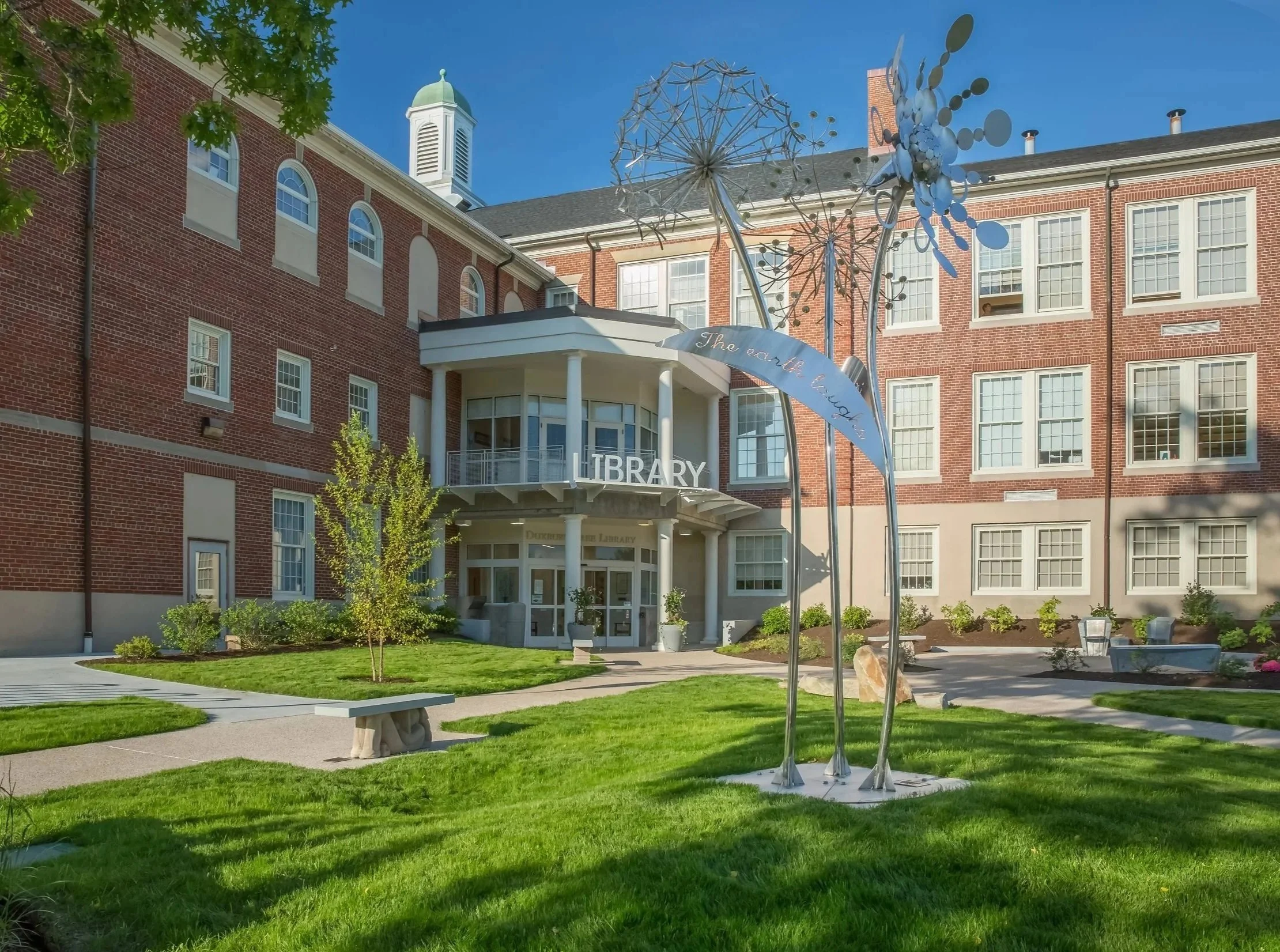 Red brick library building with white accents and a prominent sign reading 'Library' at the front entrance. The scene includes a well-maintained lawn, trees, and decorative metal artwork resembling dandelions in front of the building. Clear blue sky in the background.