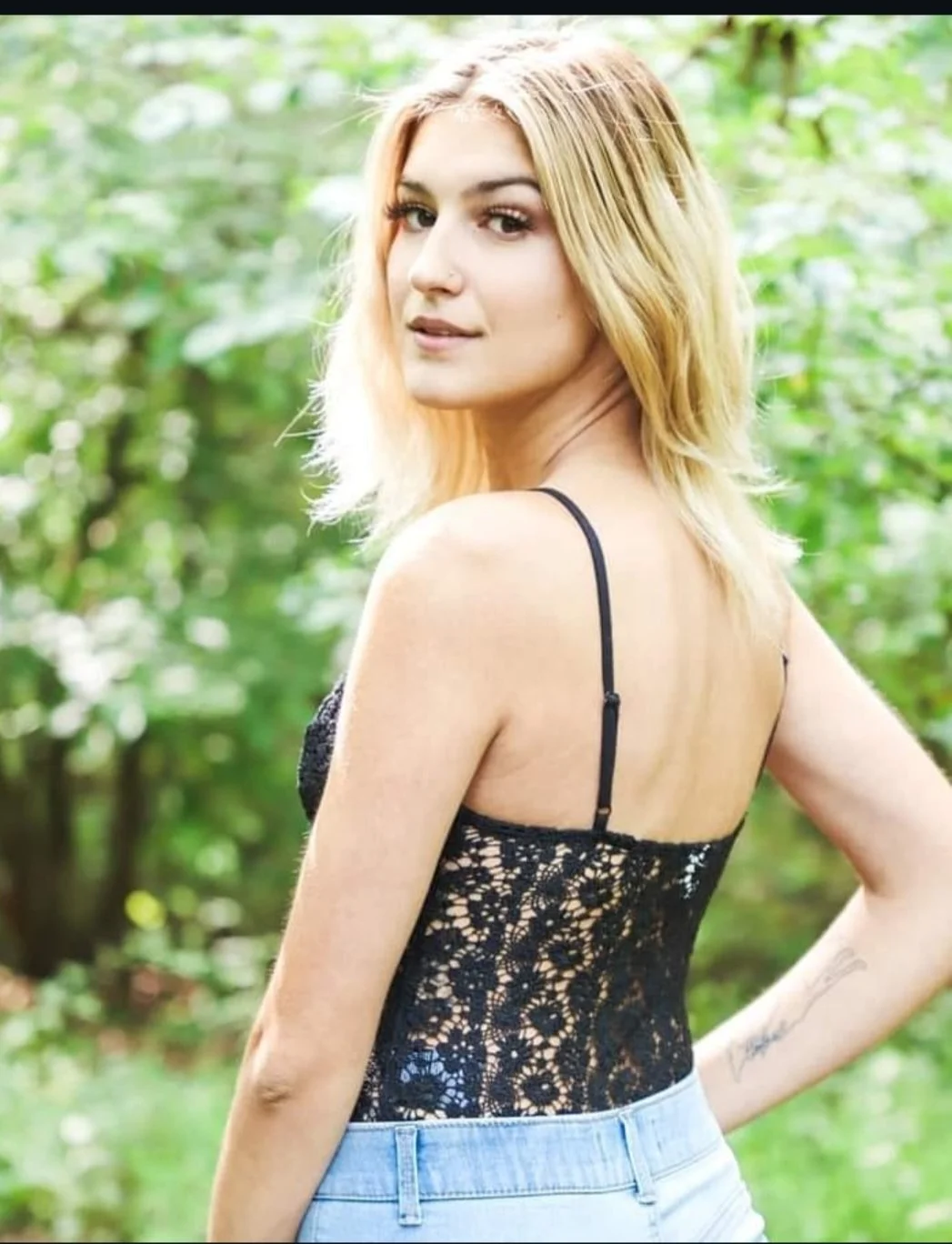 A young woman with blonde hair in a black lace top and light blue jeans standing outdoors in a green, leafy area.