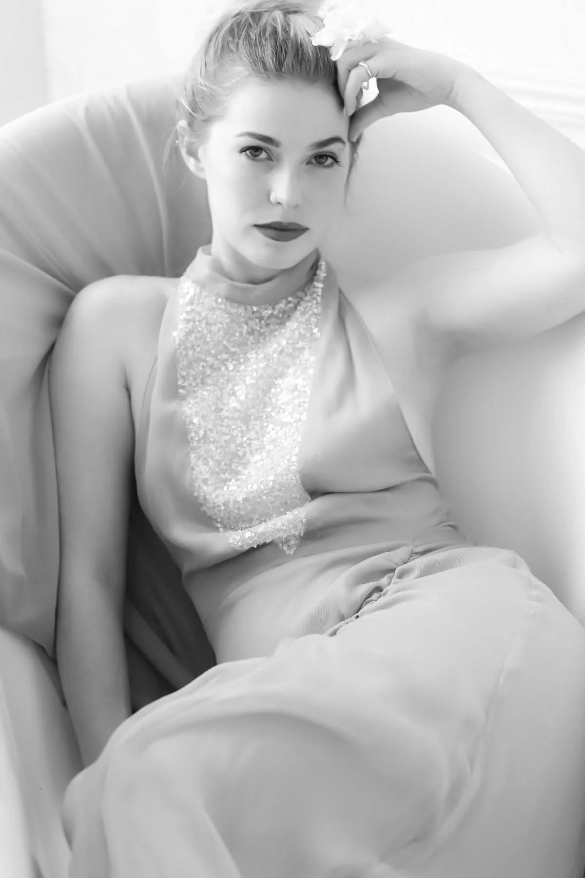 Black and white photo of a woman sitting on a sofa, wearing a sleeveless dress with a sequined front, with one hand resting on her forehead and the other on her lap, looking directly at the camera.