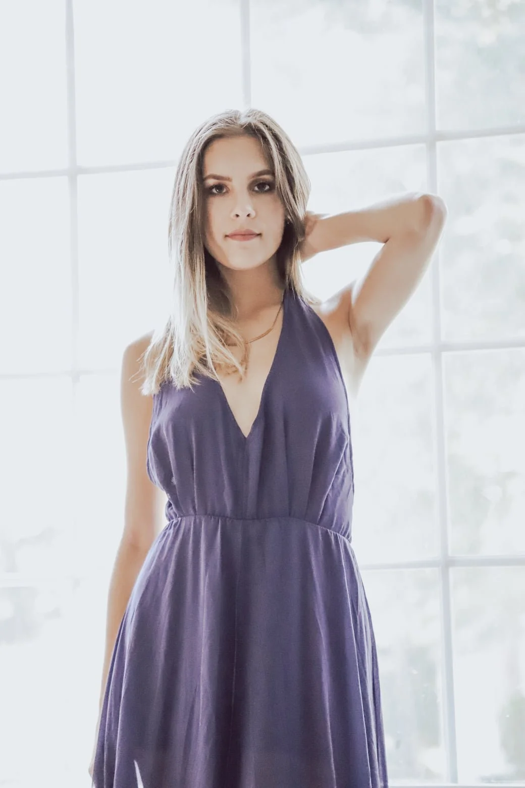 Young woman with shoulder-length brown hair, wearing a dark purple sleeveless dress, standing in front of a large window with white grid panes, smiling softly and looking at the camera.
