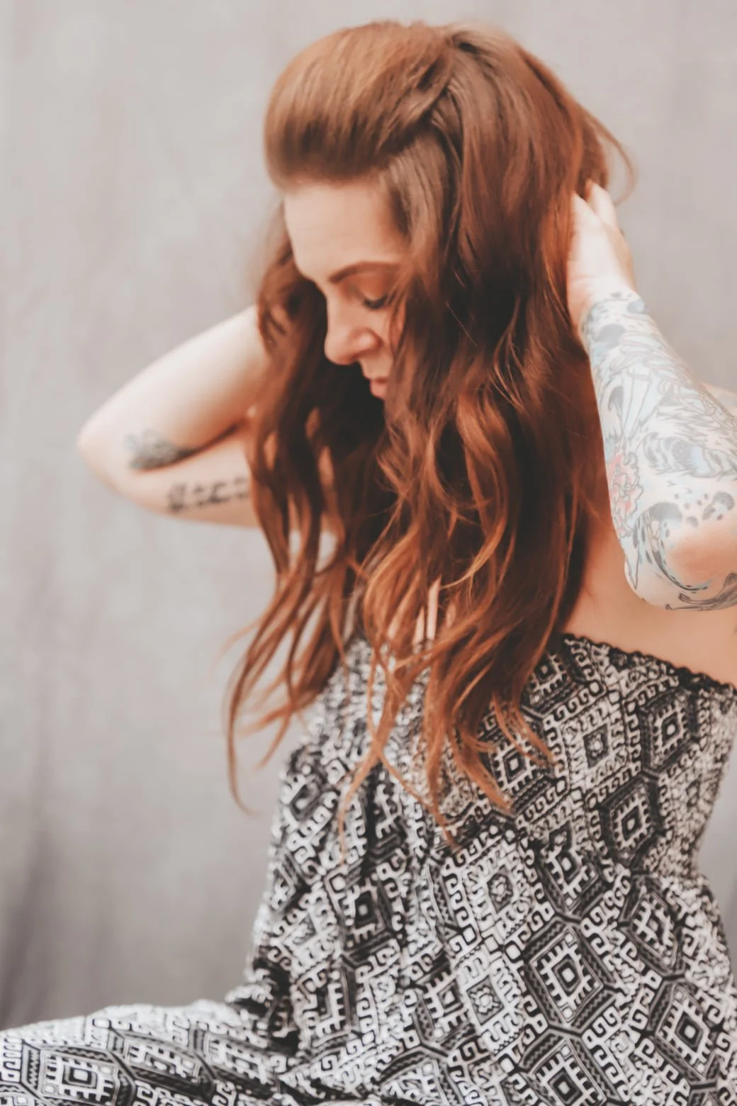 A woman with long, wavy red hair, tattoos on her arms, wearing a patterned skirt, posing against a plain background with her eyes closed and head tilted down, touching her hair.