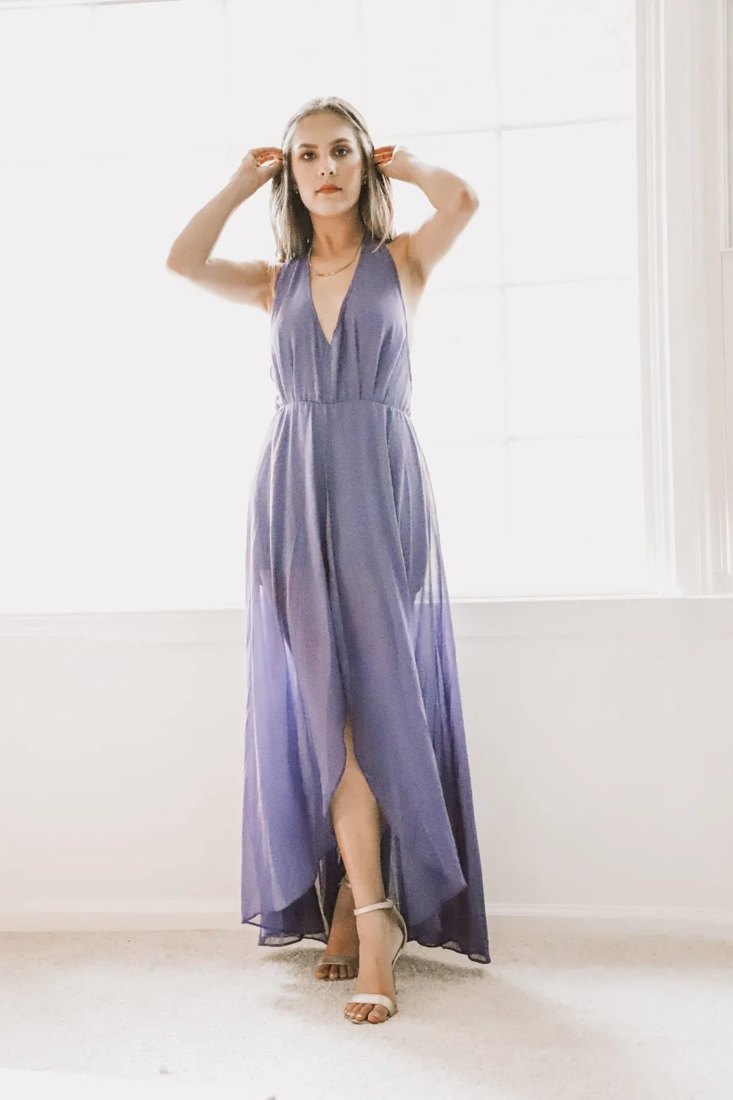 A woman in a purple, sleeveless, V-neck gown with a slit, standing on a carpeted floor in front of a bright window, wearing white high heels.