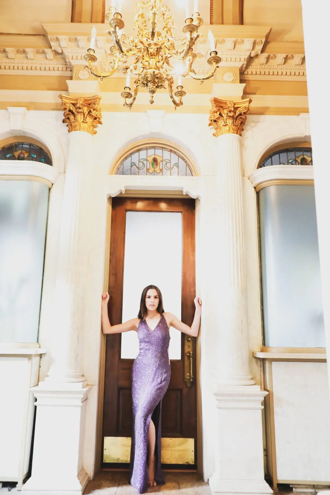 Woman in a shimmering purple gown standing in front of a large wooden door with ornate white columns and stained glass windows, a chandelier hanging above.