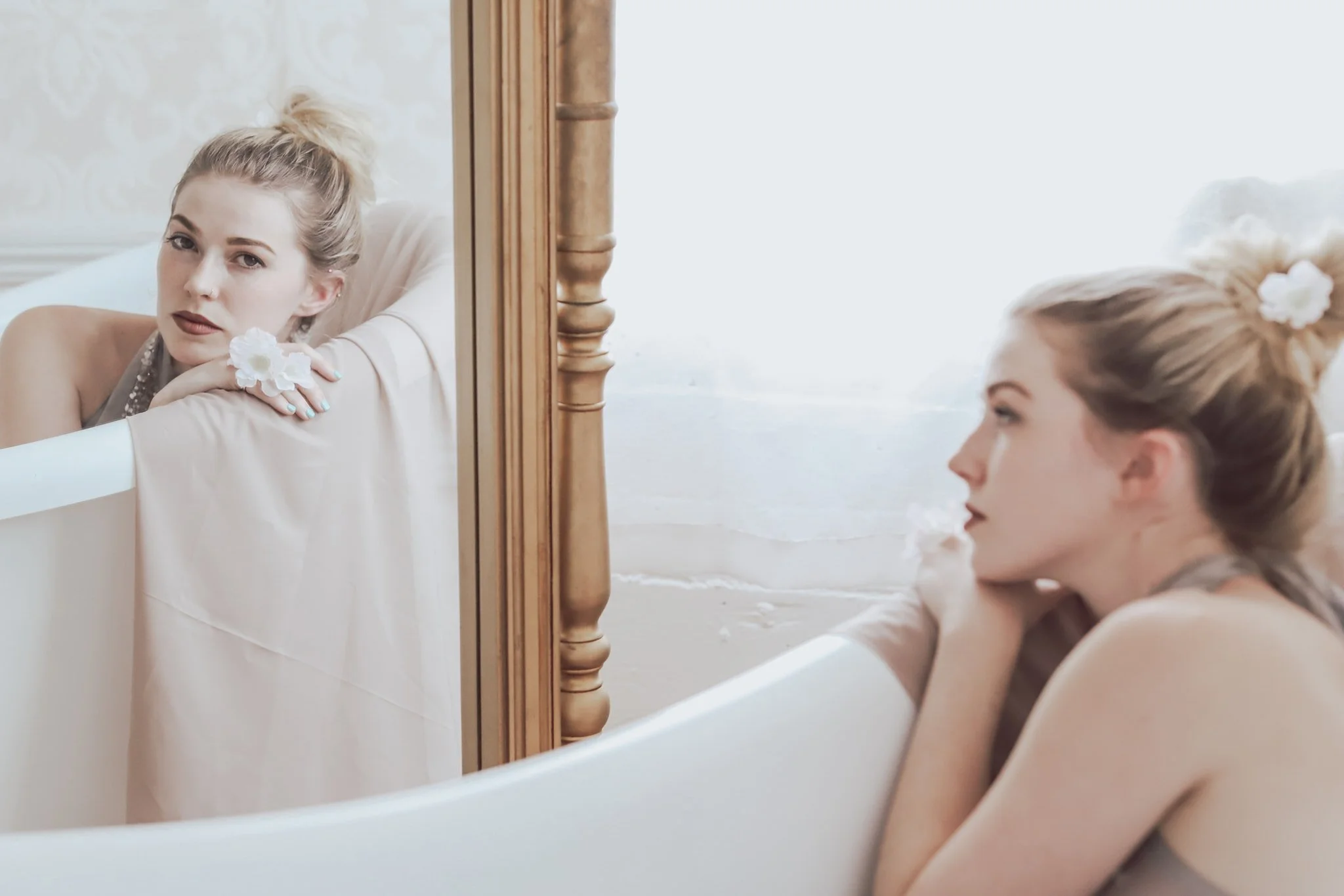 A woman with blonde hair in a bun decorated with a white flower, sitting by a bathtub, looking at her reflection in a mirror.