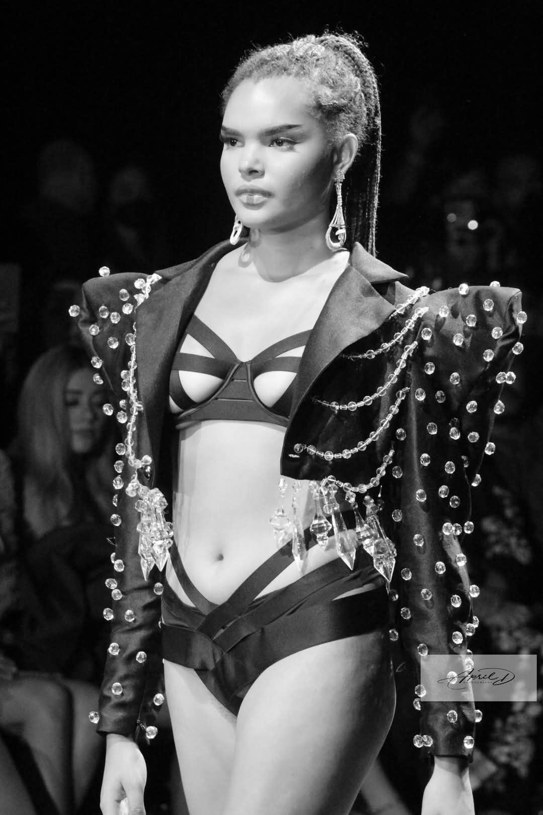 A female model walking on a runway during a fashion show, wearing a black outfit with a jacket decorated with chains and beads, a strappy bikini top, and high-waisted bottoms, with her hair in braids and wearing earrings.