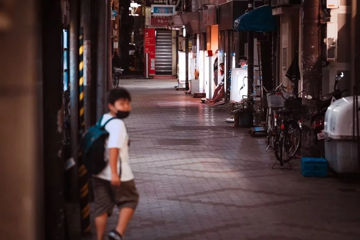 A young boy wearing a face mask, white shirt, and shorts walking on a dimly lit city sidewalk at night, with storefronts and bicycles along the side on the streets of Osaka.