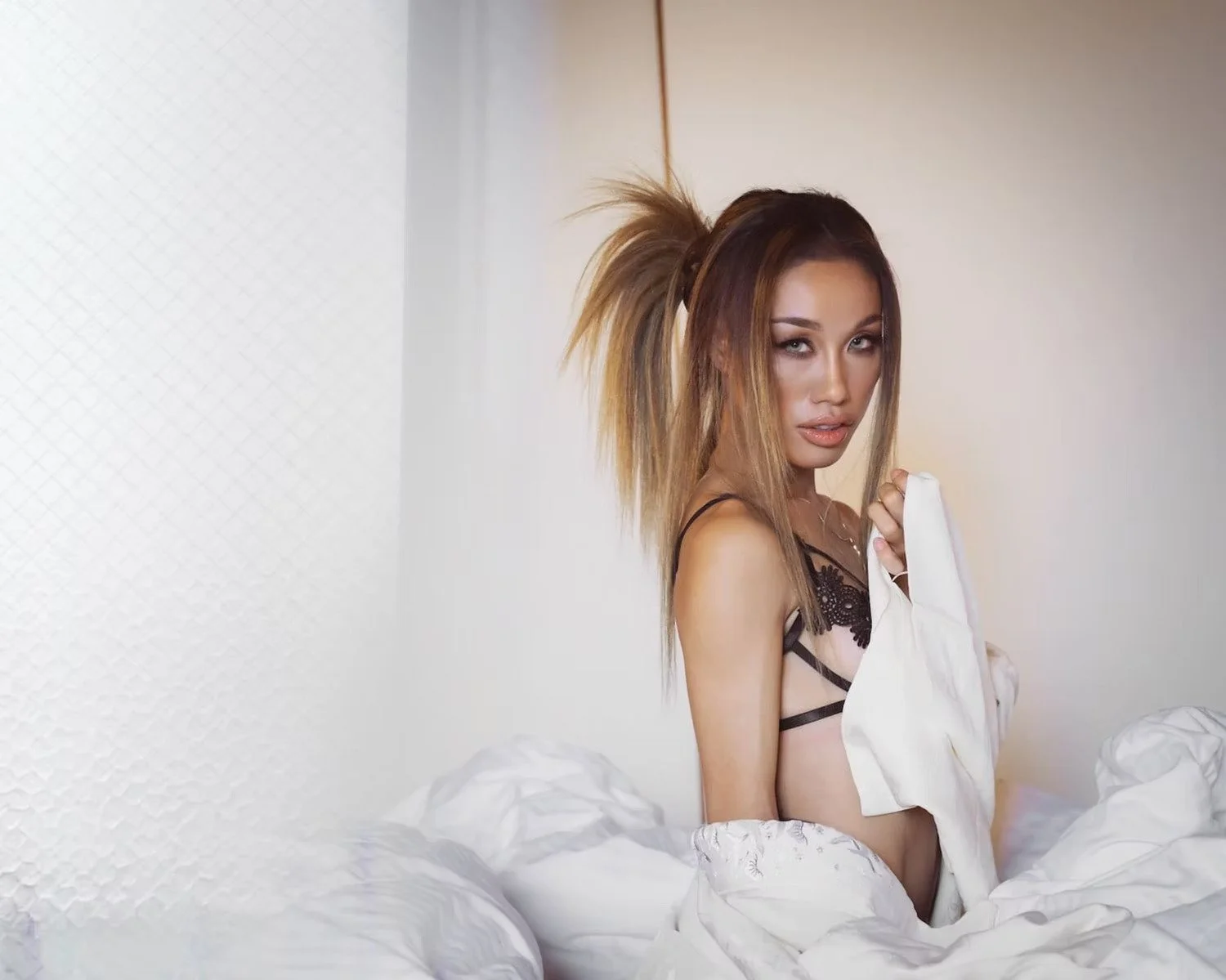 A woman with long brown hair styled in a high ponytail and wearing black lingerie posing in bed with white bedding, holding a white shirt.