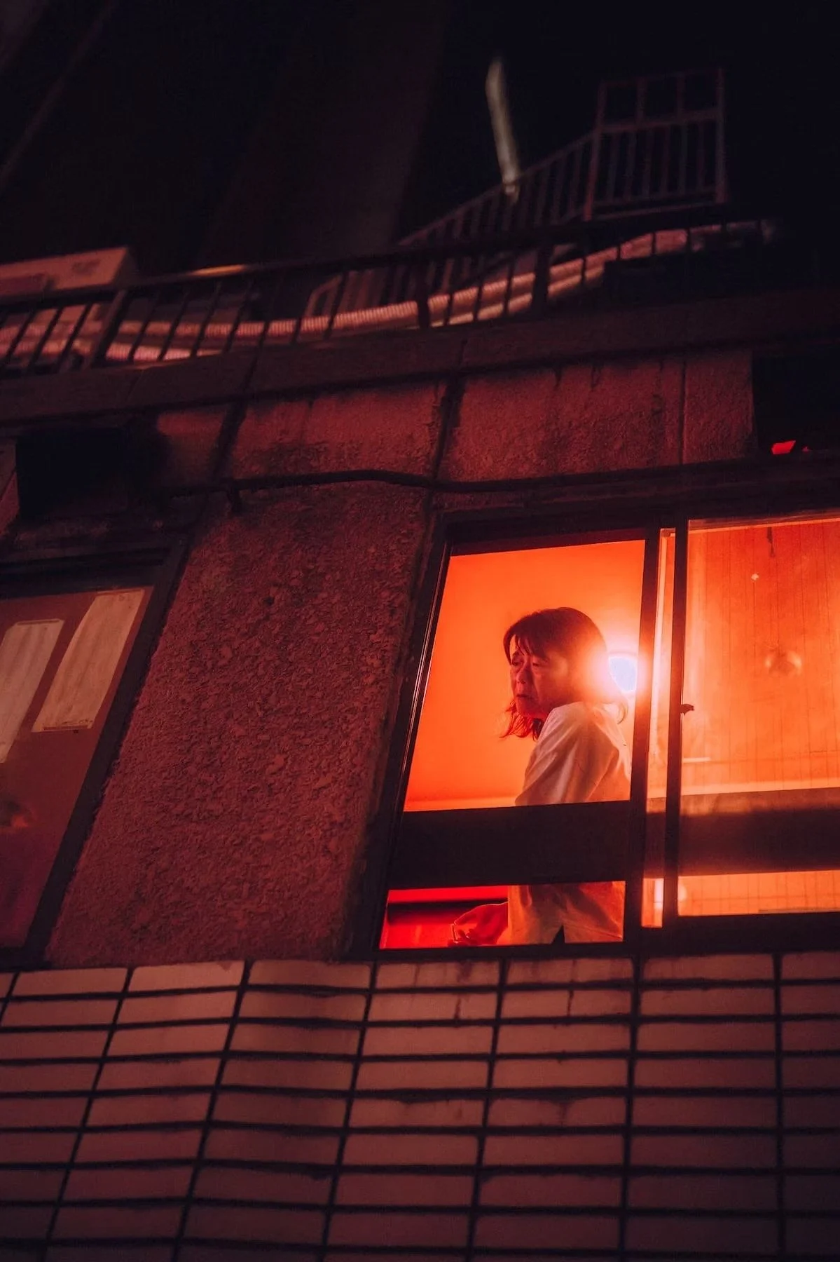 A Japanese woman with medium-length hair stands inside a warmly lit room, looking out of a window at night. The room has an orange-red glow, and she appears to be lost in thought.