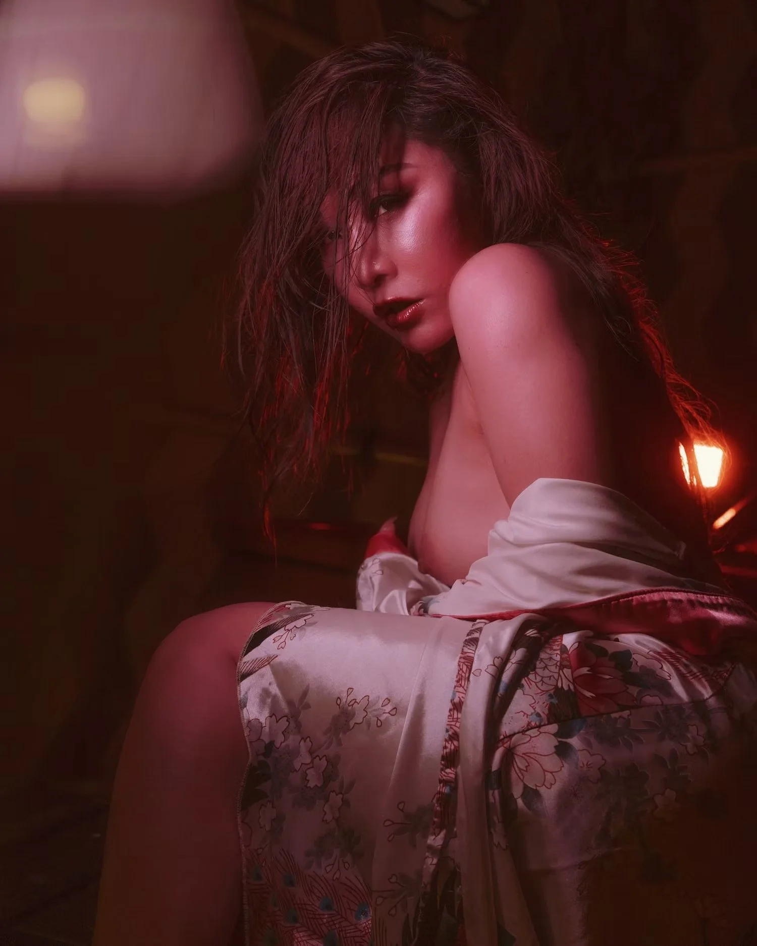 A woman with wet hair looking over her shoulder in a dimly lit room, wearing a silk garment with floral patterns.