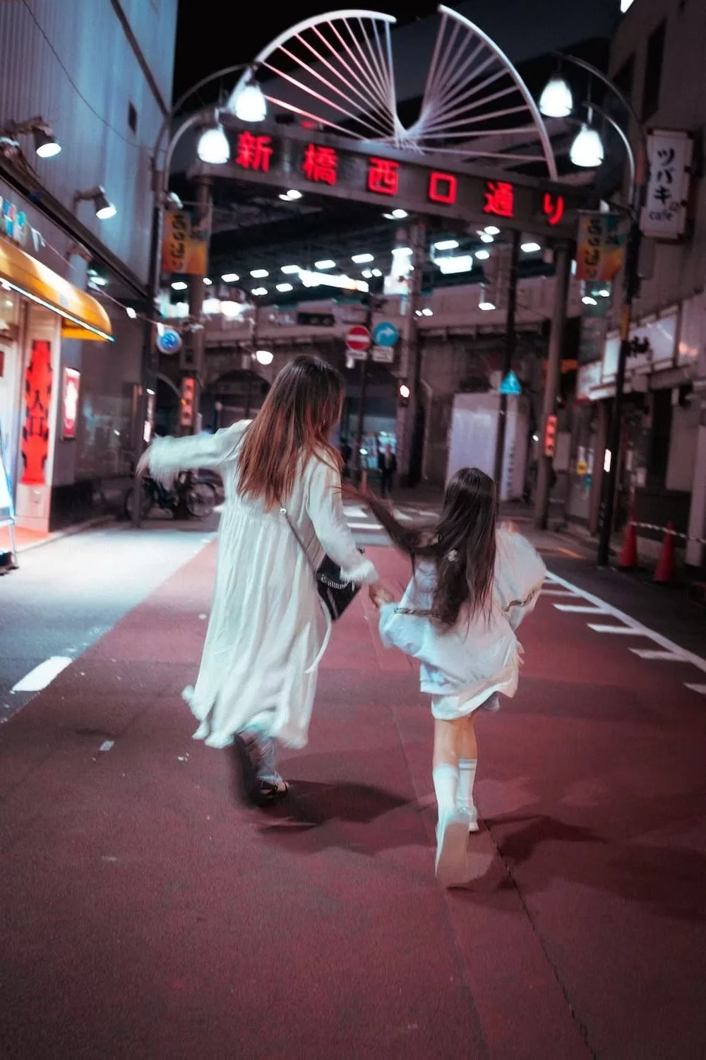 A mother and child in white skirts and long coats running on a city street at night with neon signs and streetlights, holding hands and their hair flowing behind them on the streets of Shimbashi Tokyo in Japan.