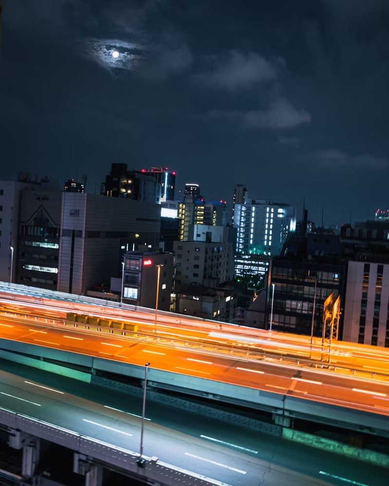 Nighttime cityscape with illuminated high-rise buildings, a bright full moon partially covered by clouds, and moving traffic on highways creating light trails.
