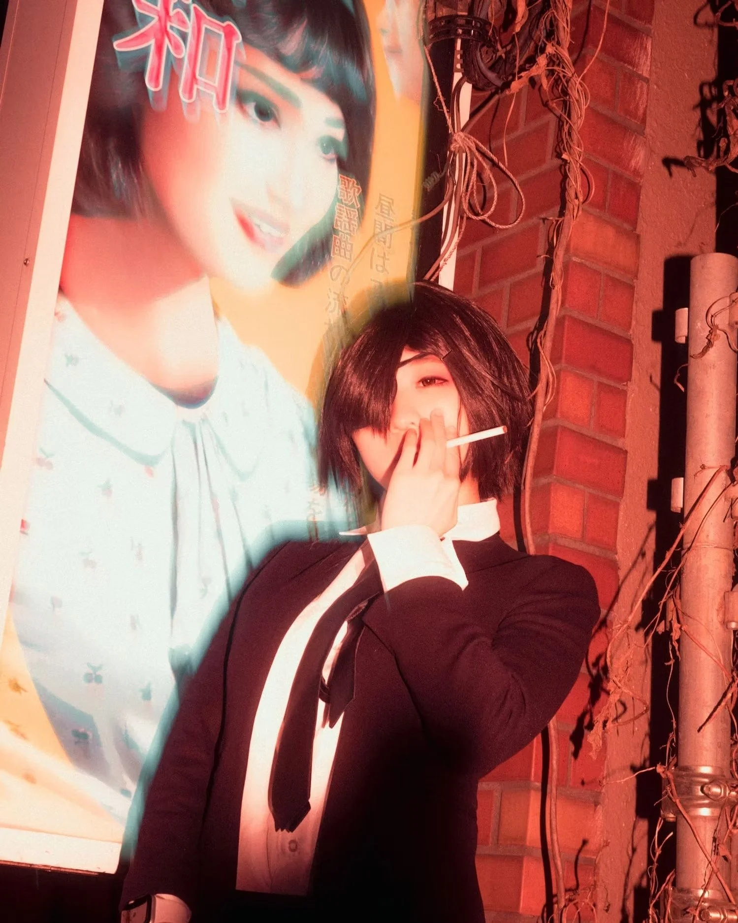 A person with short dark hair, wearing a black outfit with a white stripe, holding a cigarette in their right hand, standing in front of a poster featuring a woman's face with short black hair, making a V sign with their left hand, against a brick wa