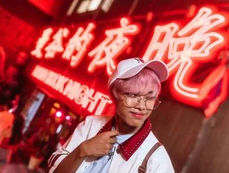 Person with pink hair, glasses, white cap, and backpack standing in front of neon red sign in an urban setting.