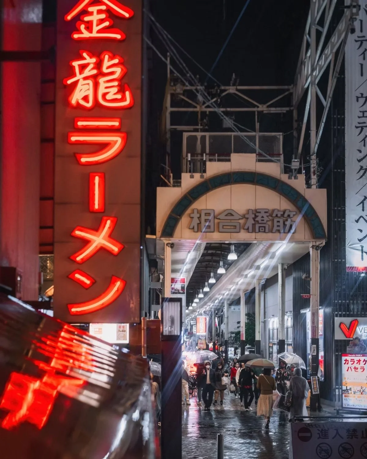 Night scene of a busy street with neon signs in Japanese, people walking with umbrellas, and a covered shopping arcade in Osaka, Namba district.
