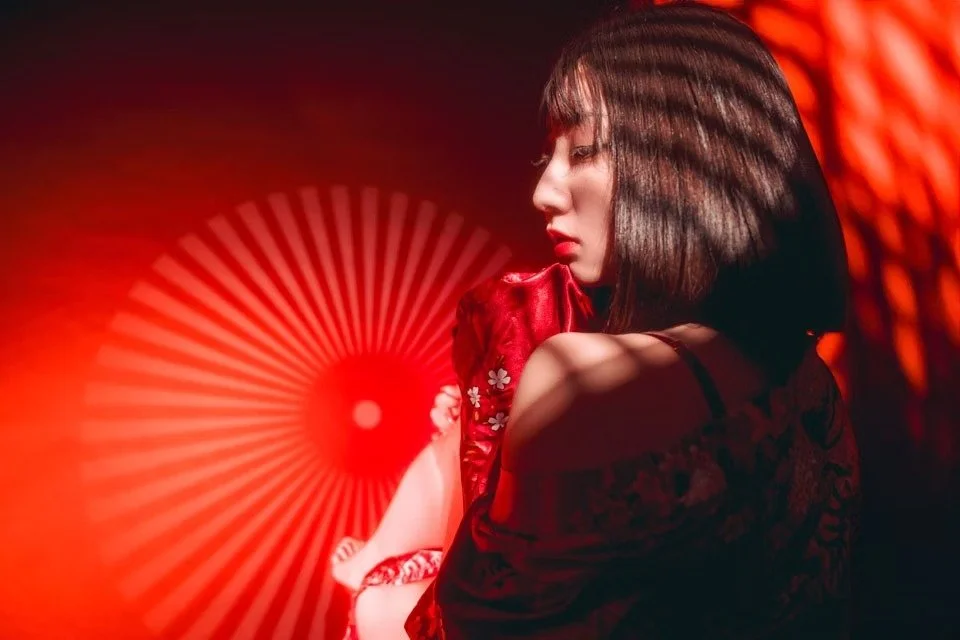 A woman with short hair holds a fan to the side of her face, surrounded by red lighting. The contrasting lighting and shadows give the image a bold, sensual vibe.