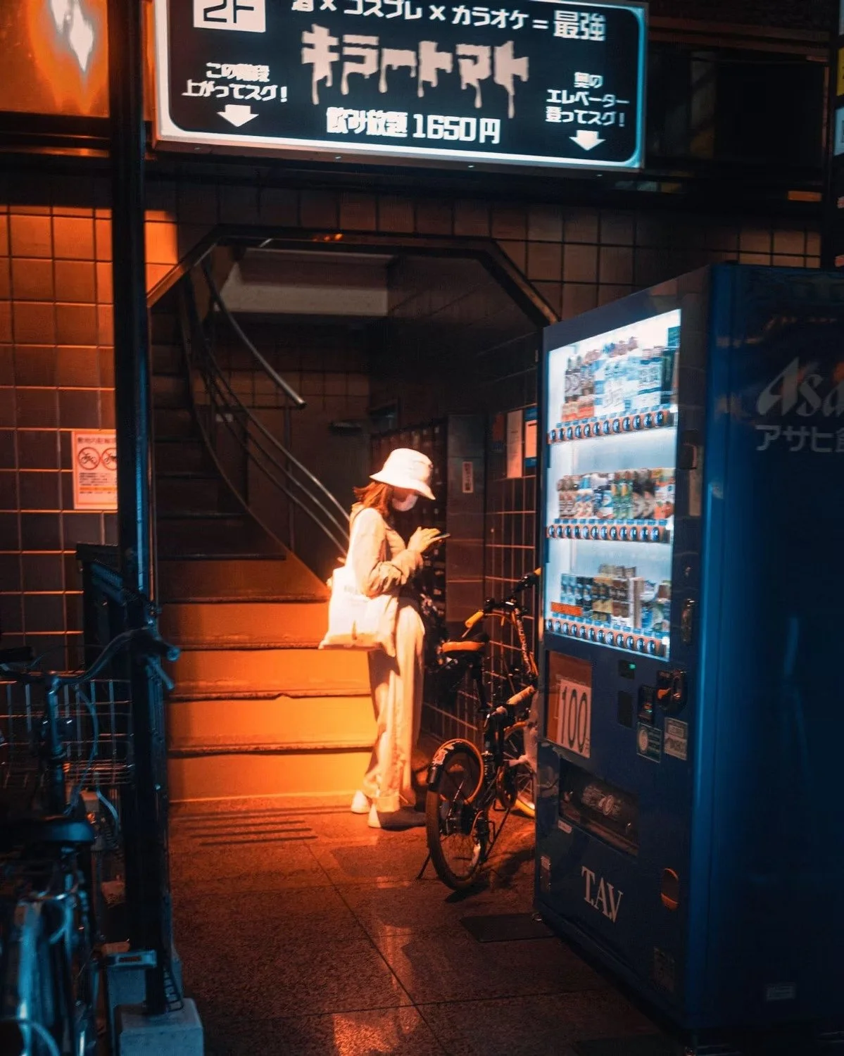 A woman wearing a hat and face mask sitting on stairs at night, looking at her phone. Bicycle parked nearby and vending machine in front. Dim orange lighting shot in Osaka.
