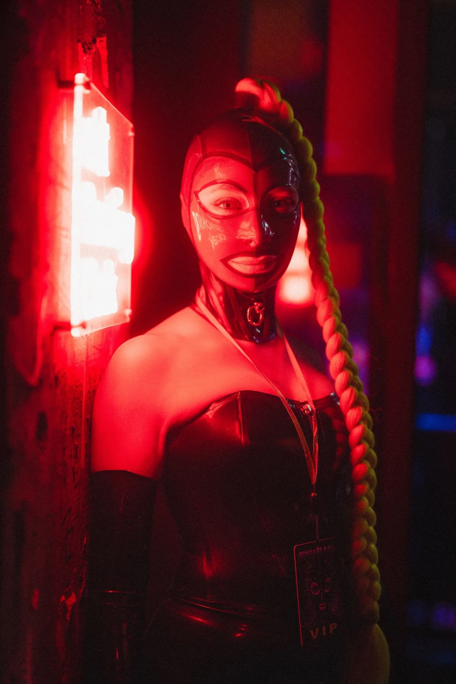 Bondage Ball 
---
A woman with light skin wearing a shiny black mask and a zipper top, illuminated by red and blue lights, with long braided hair, standing in a dark room.