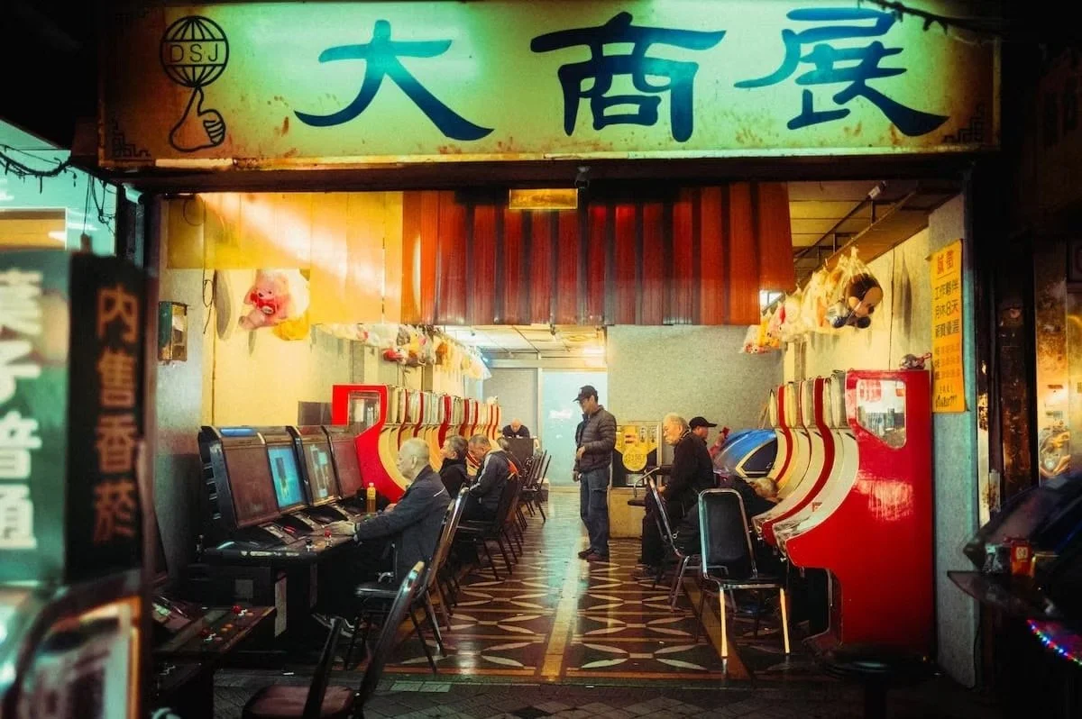 Interior of an arcade with elderly people playing on slot machines, a man stands in the center, and colorful machines line the walls under a sign with Chinese characters. Retro Taiwan captured by street photographer Deniz Demir.