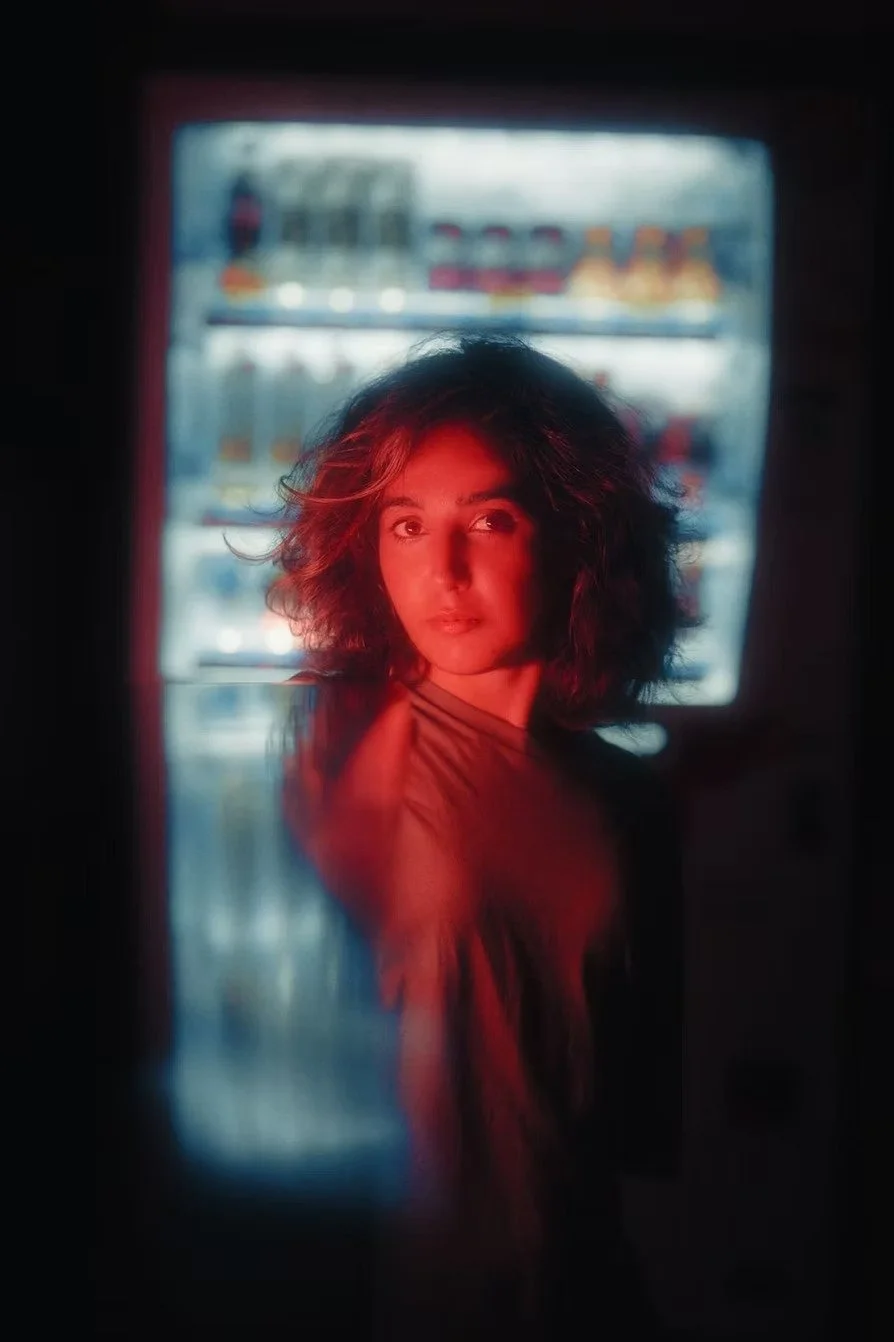 A woman with curly hair looking through a vending machine with a red light illuminating her face.