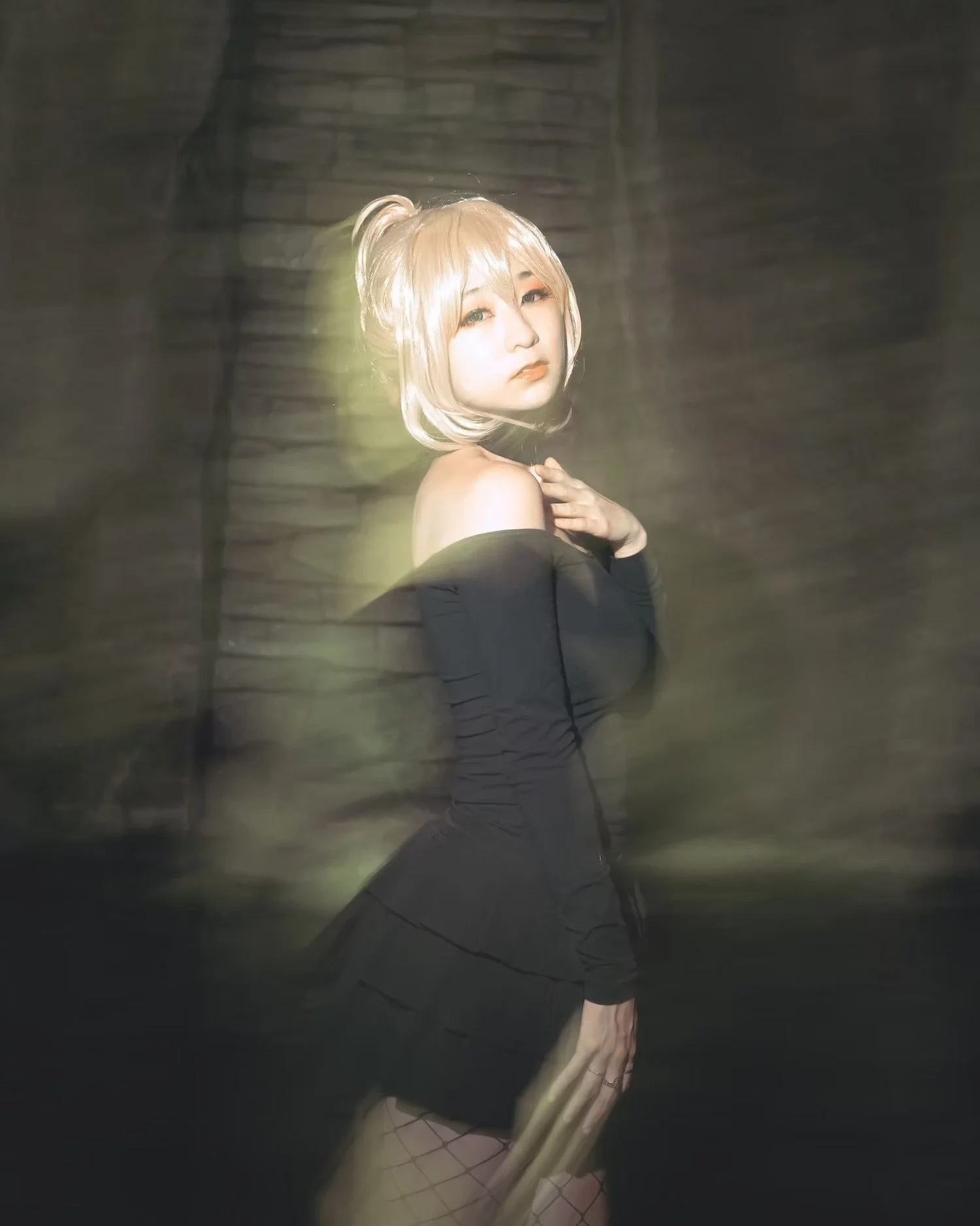 A woman with blonde hair styled in a high ponytail, wearing an off-the-shoulder black dress and fishnet stockings, poses in a dimly lit setting with blurry dark background.