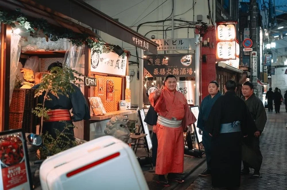 Group of Japanese Sumo wrestlers in Koenji in traditional kimonos standing outside a restaurant on a busy street, with one man in a red kimono making a peace sign.