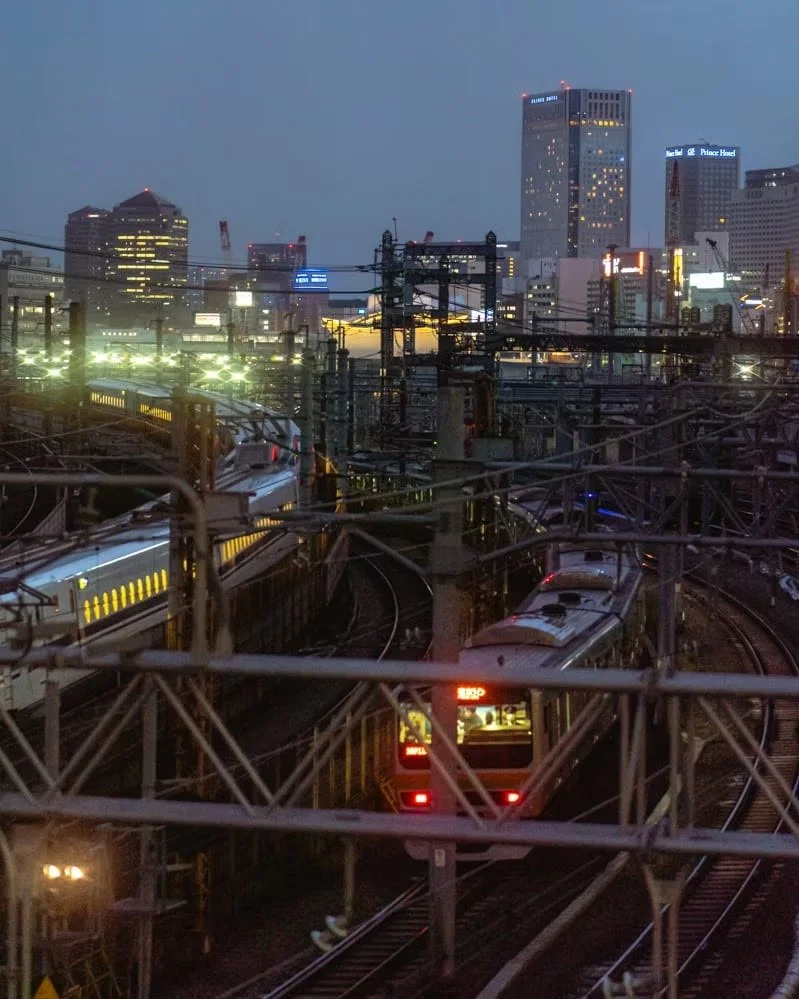 A train traveling through a city at dusk with tall skyscrapers and numerous overhead power lines.