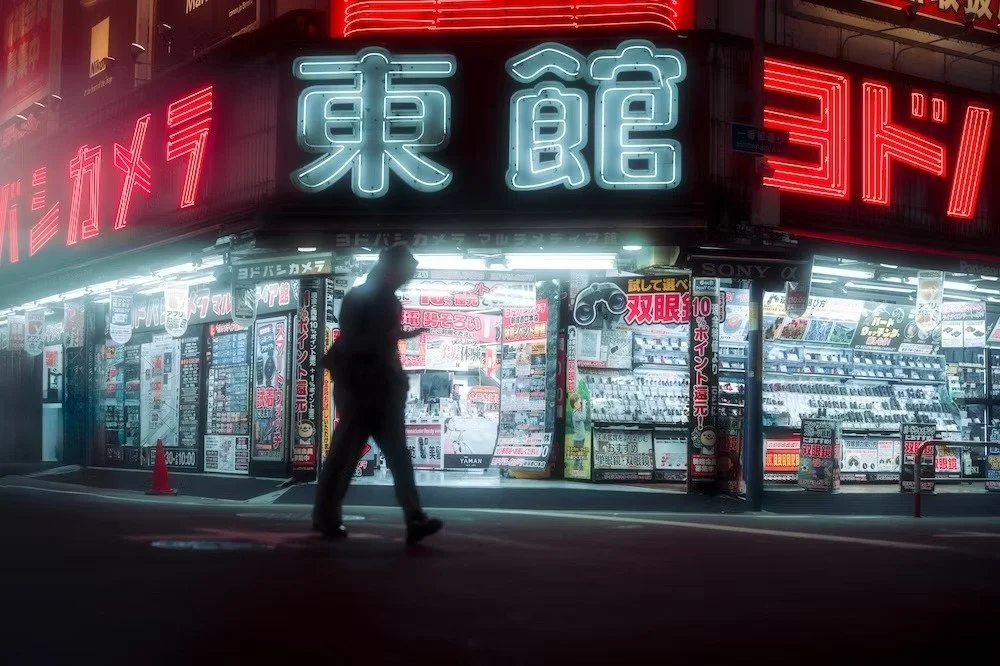 A person walking in front of a brightly lit Japanese electronics store at night, with neon signs and displays of cameras and electronic products shot in Shinjuku Yodobashi area by Tokyo street photographer Deniz Demir.
