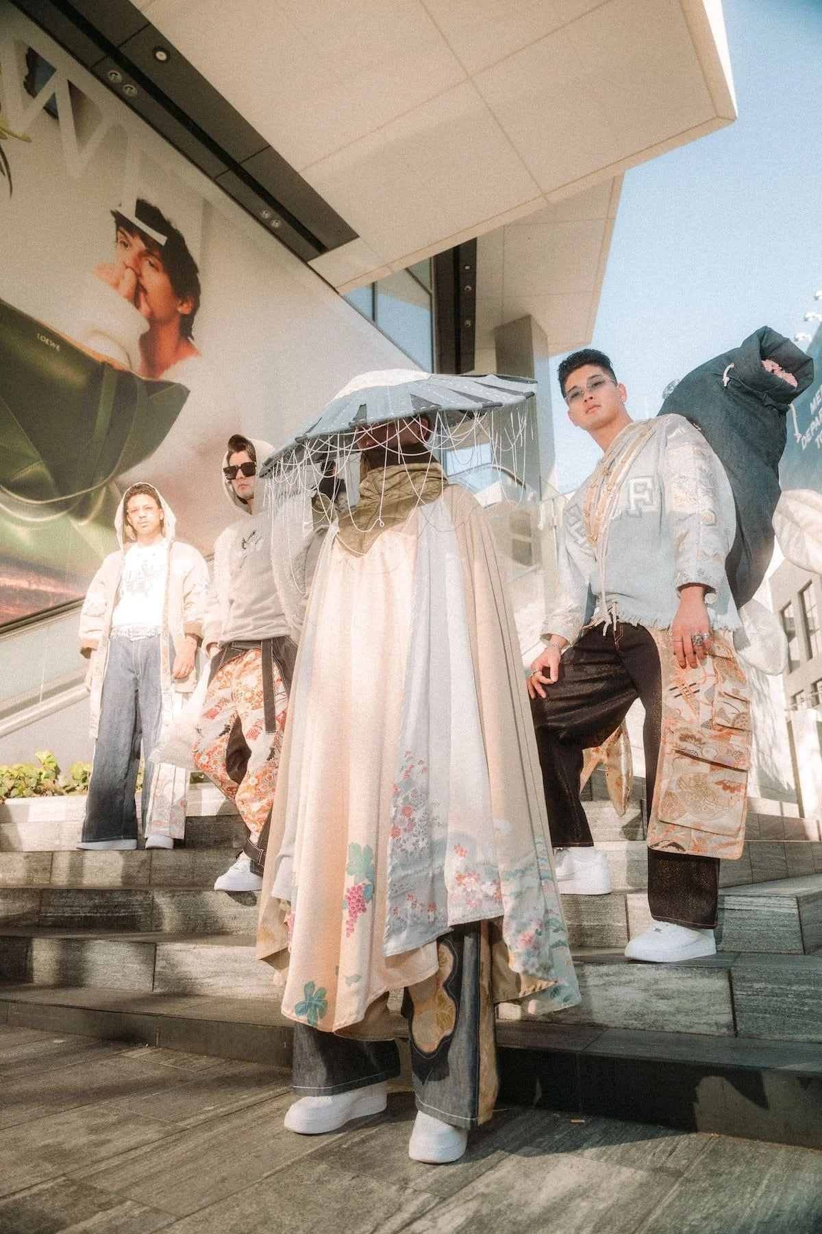 A group of five young people standing on steps outside a modern building, dressed in casual and streetwear clothing, with one person holding an umbrella and wearing traditional Asian attire, while a large advertisement featuring a woman appears on the building behind them.