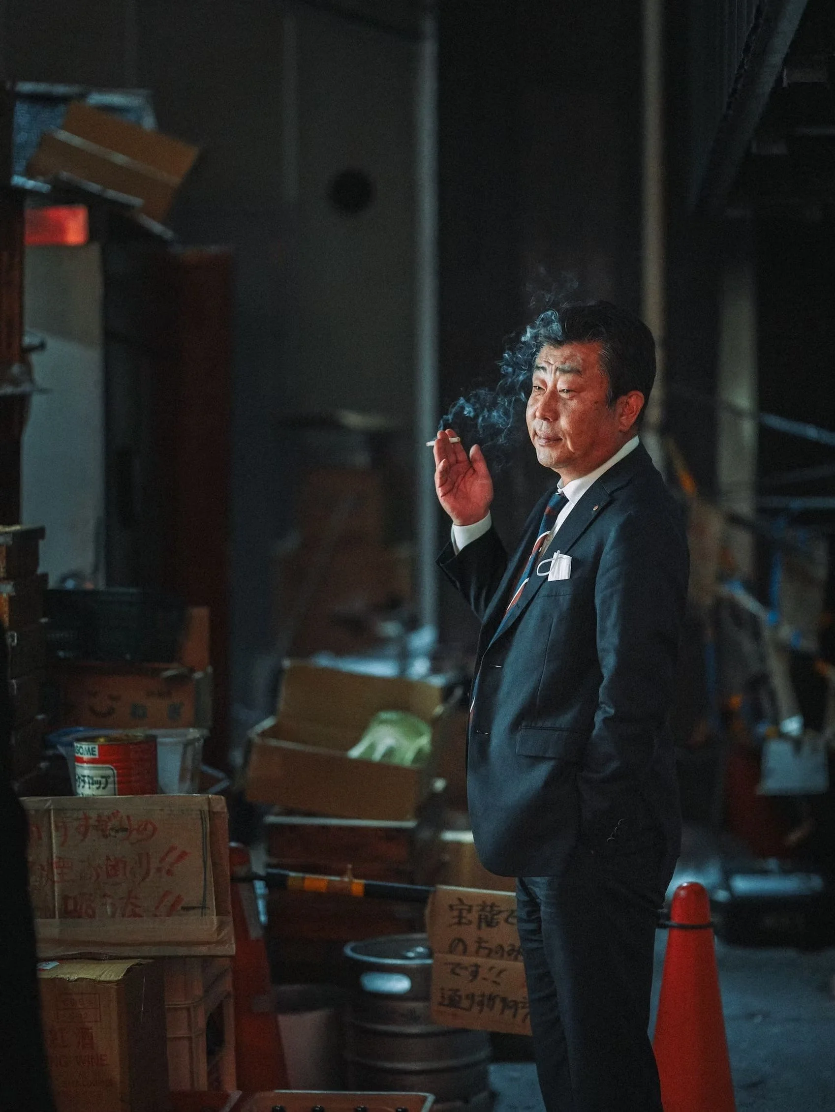 A man in a suit and tie, smoking a cigarette, standing in a dimly lit storage or warehouse area surrounded by boxes and shelves.