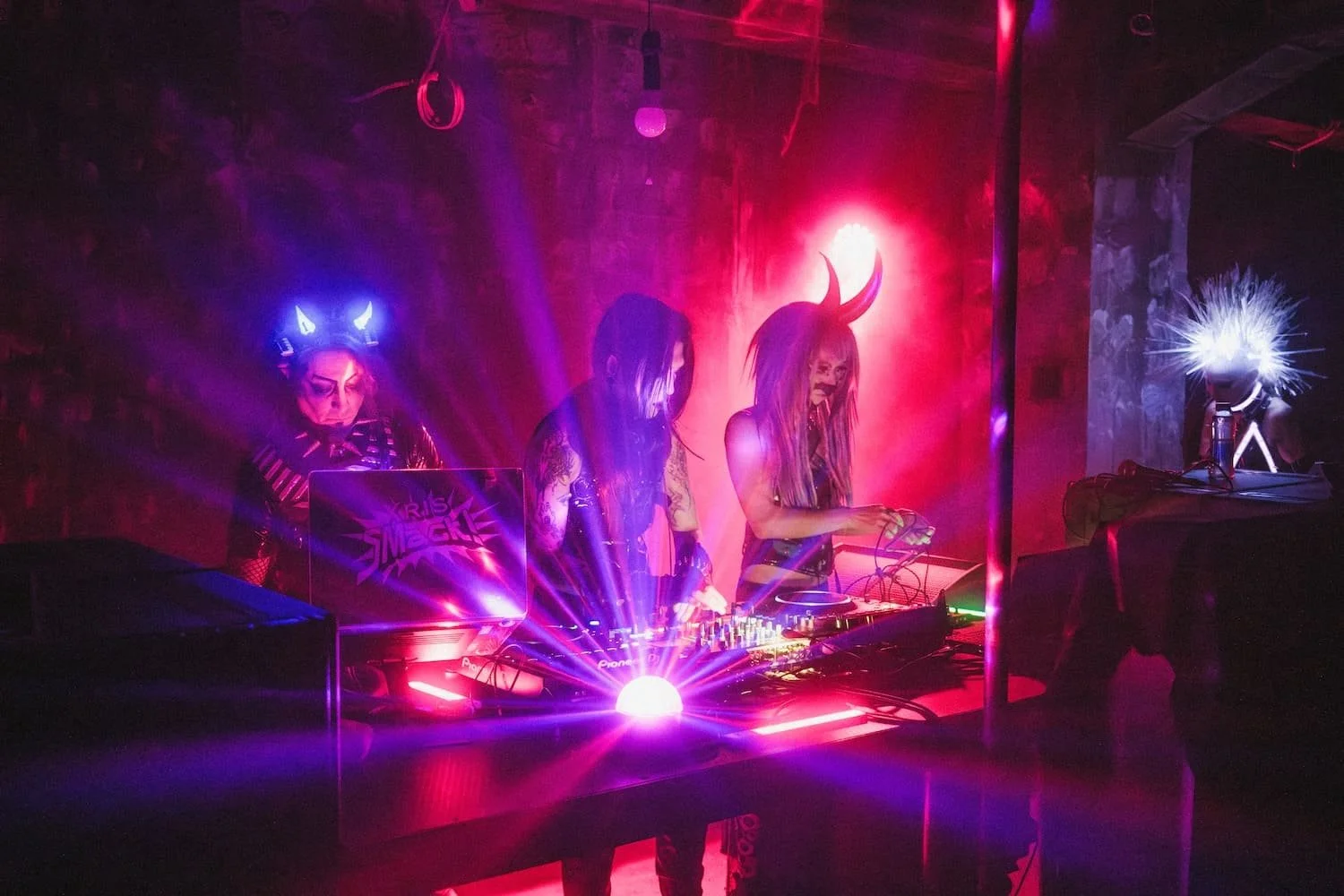 Bondage Ball 
---
Three DJs wearing Halloween costumes with neon lighting at a dark club, with one DJ in a devil costume with horns, another in a bat-themed costume with glowing ears, and a third with spiked hair and face paint, surrounded by colorfu